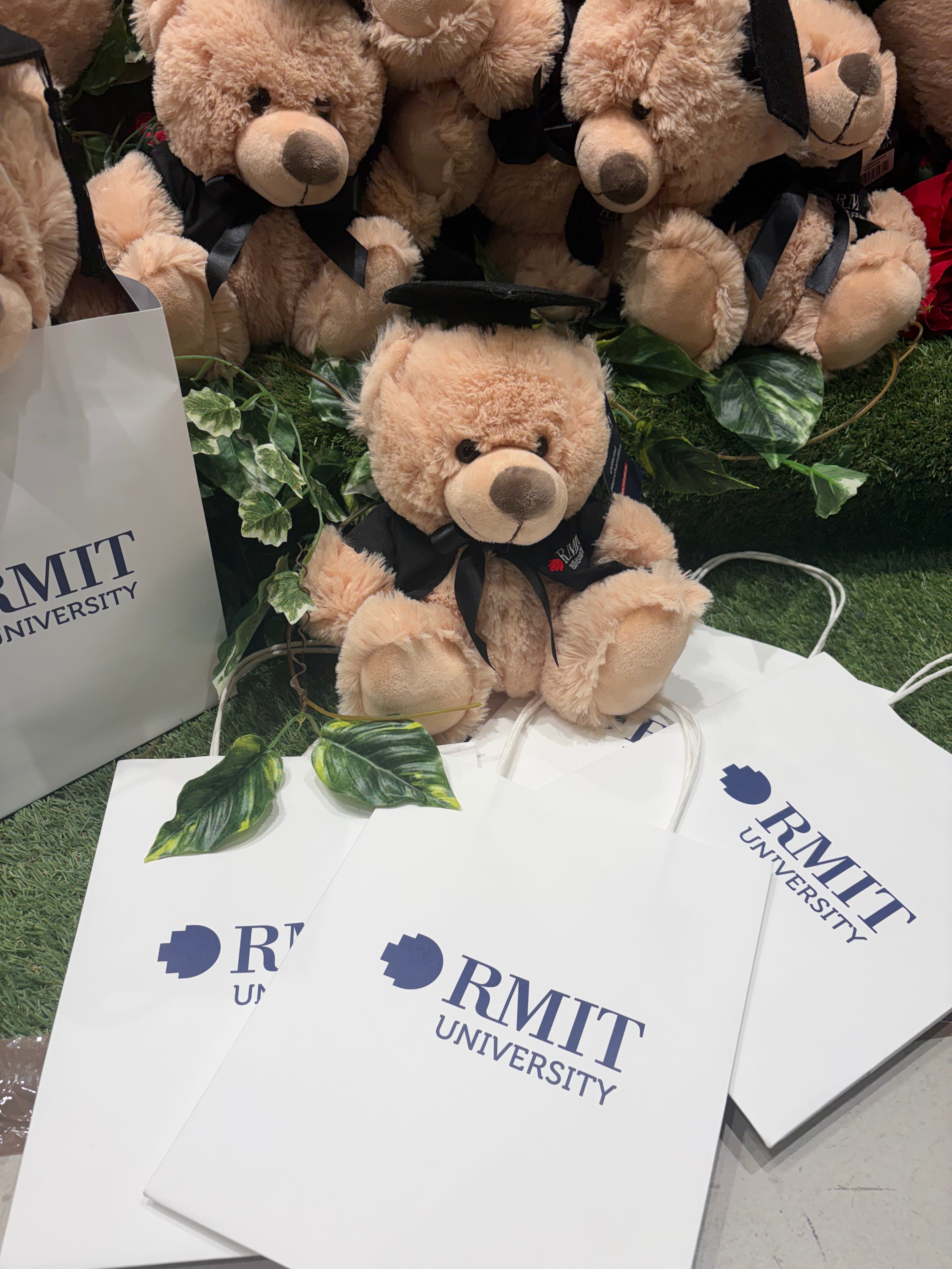Multiple white RMIT University gift bags laid flat on artificial green grass with a graduation teddy bear sitting upright above them, surrounded by additional plush bears and greenery inside the RMIT Store.