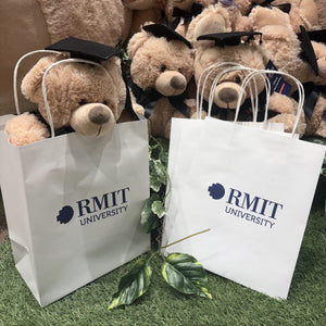 Two white RMIT University gift bags with twisted handles standing on artificial grass in front of a display of graduation teddy bears wearing black mortarboards inside the RMIT Store.