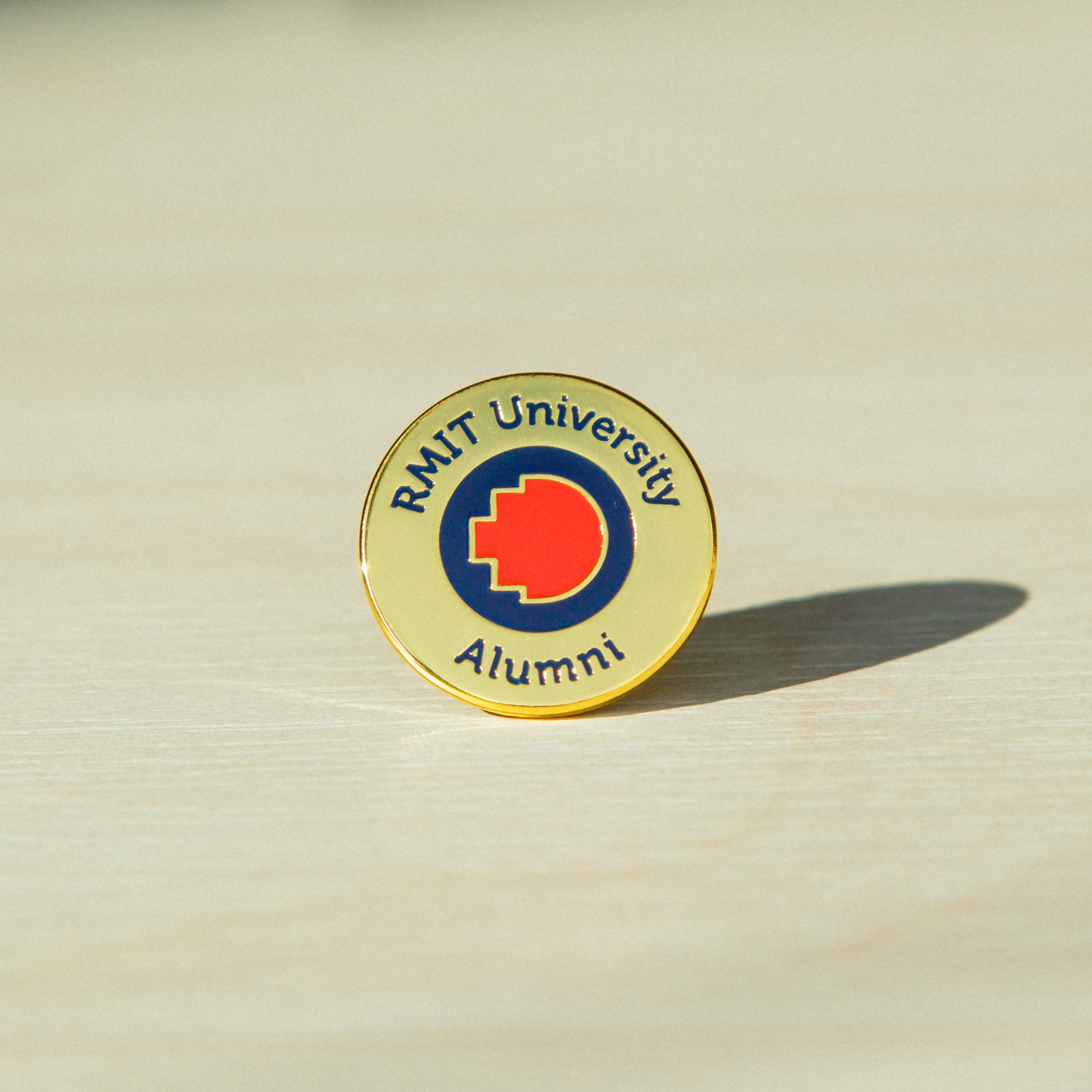 Close-up of a gold metal RMIT alumni pin standing upright on a wooden tabletop. The pin features an engraved “RMIT University Alumni” text around the edge of the round pin, with the RMIT logo prominently centred. The polished gold surface reflects soft natural light.