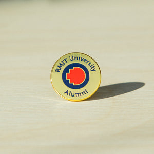 Close-up of a gold metal RMIT alumni pin standing upright on a wooden tabletop. The pin features an engraved “RMIT University Alumni” text around the edge of the round pin, with the RMIT logo prominently centred. The polished gold surface reflects soft natural light.
