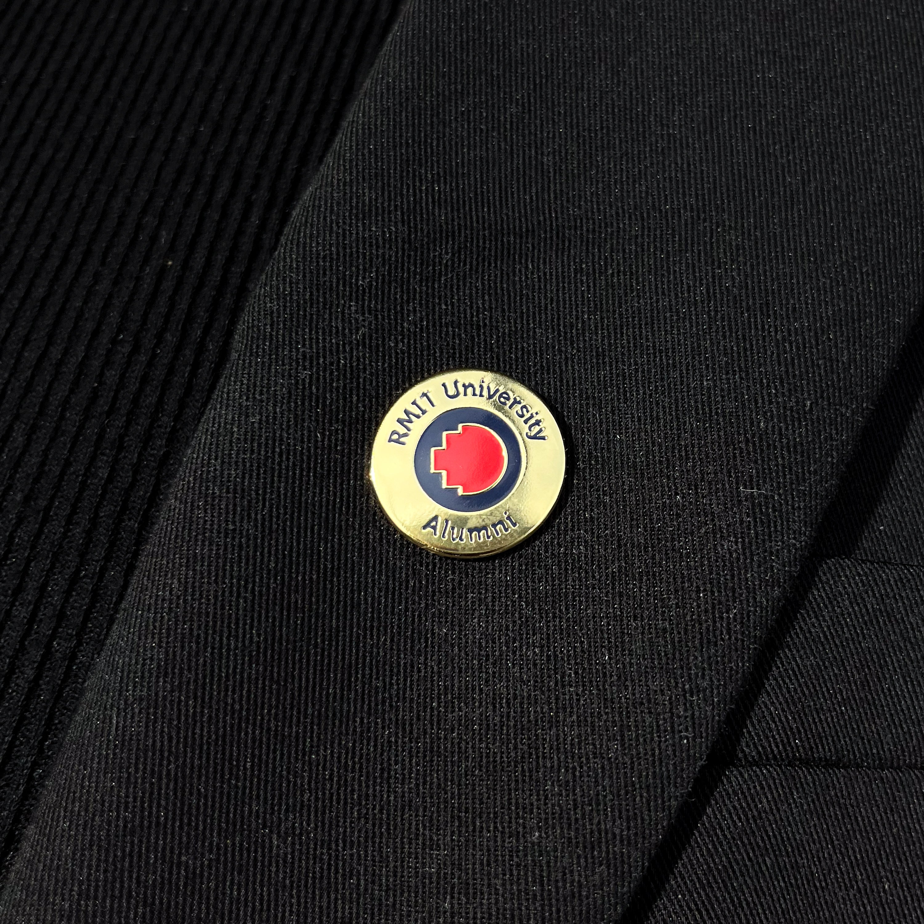 Close-up of a gold metal RMIT alumni pin attached to the lapel of a black blazer. The pin features a subtle embossed RMIT logo and a polished finish, shown against the fabric texture of the blazer.