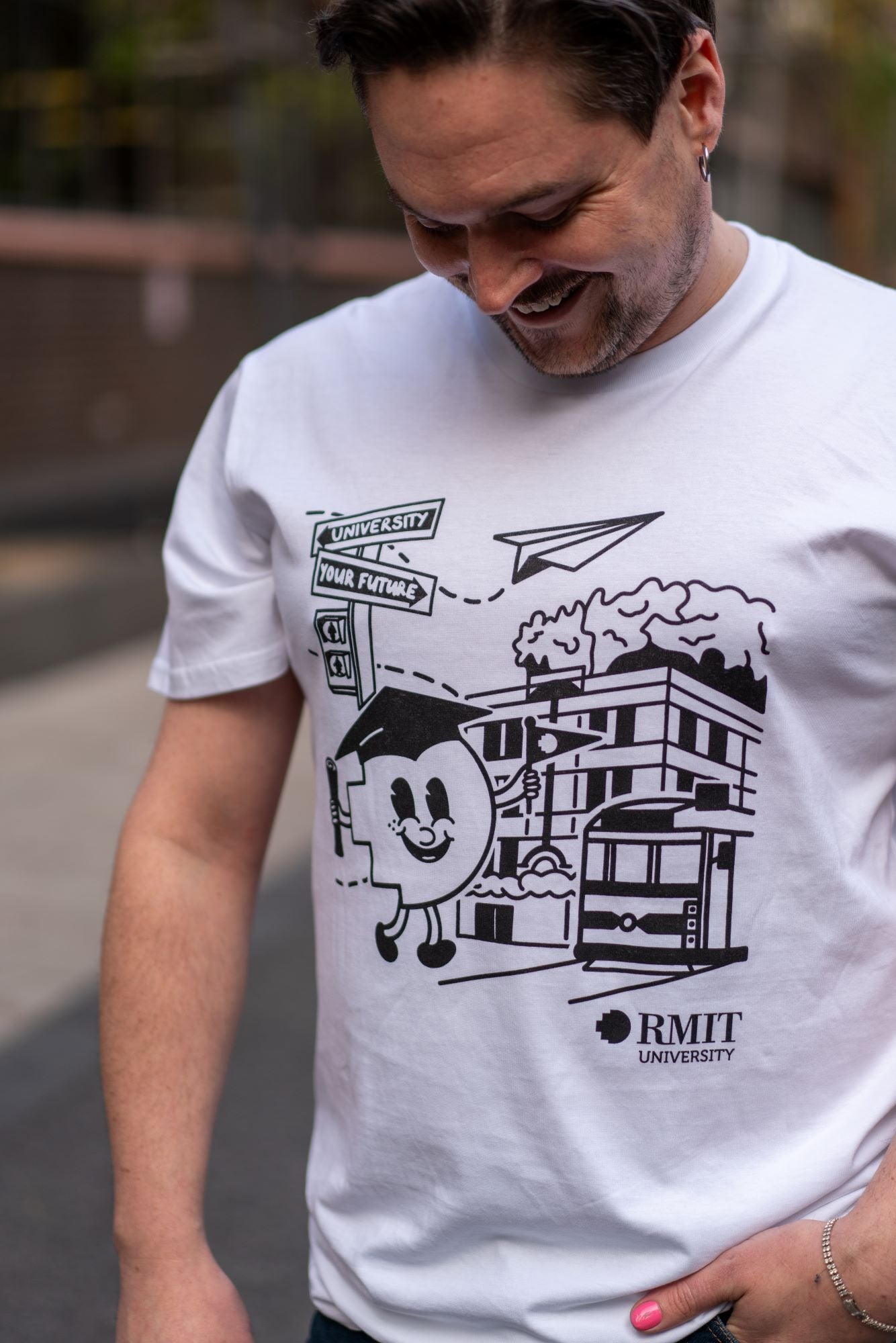 Front view of the RMIT Graduate Parade T-shirt worn by a model looking down at the black print on his chest. The white tee features an animated pixel figure in black wearing a mortarboard, positioned in front of the RMIT building on Swanston Street, available at the RMIT Store.