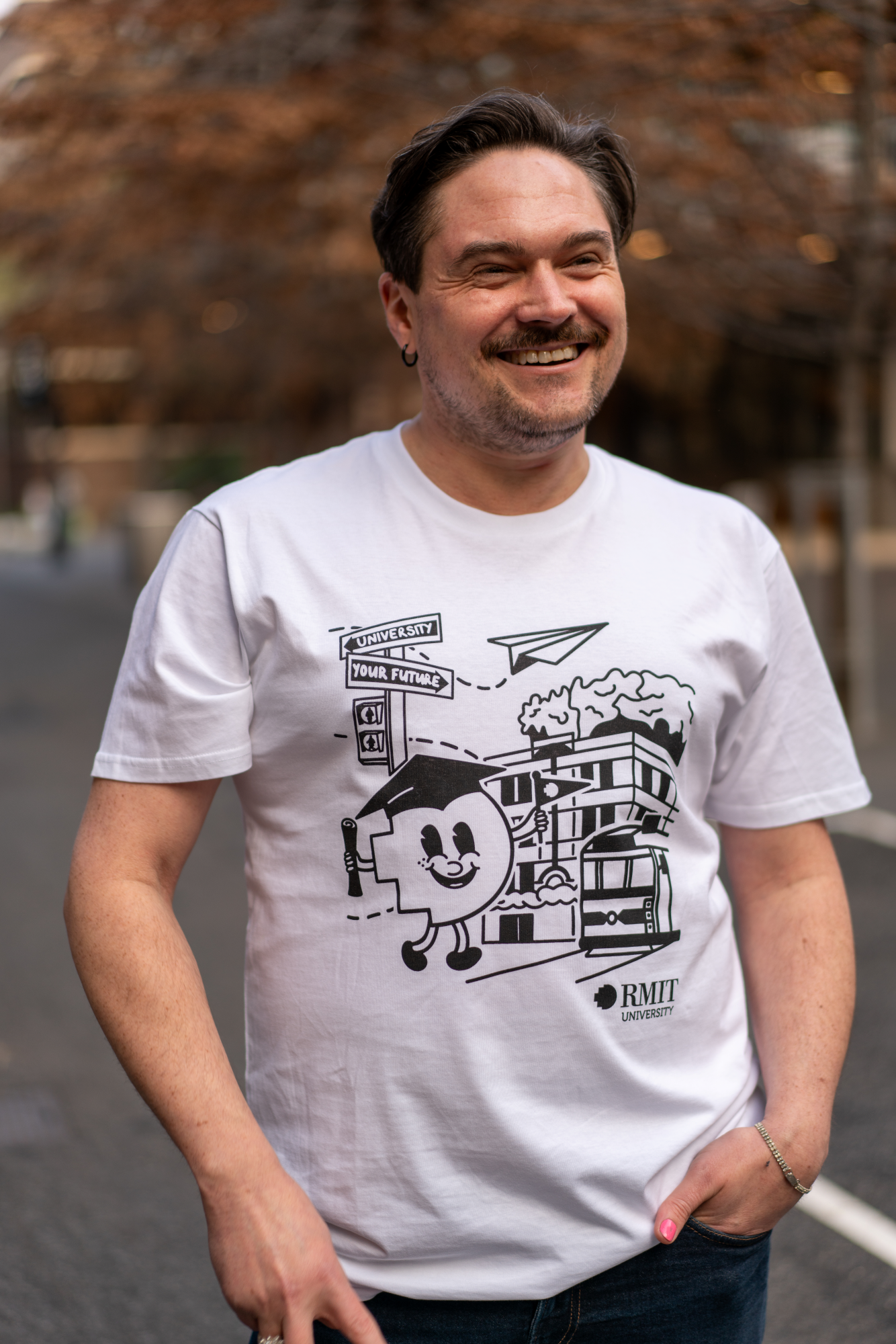 Front view of the RMIT Graduate Parade T-shirt worn by a smiling model facing the camera. The white tee features a black print of an animated pixel figure wearing a mortarboard, standing in front of the RMIT building on Swanston Street, available at the RMIT Store.