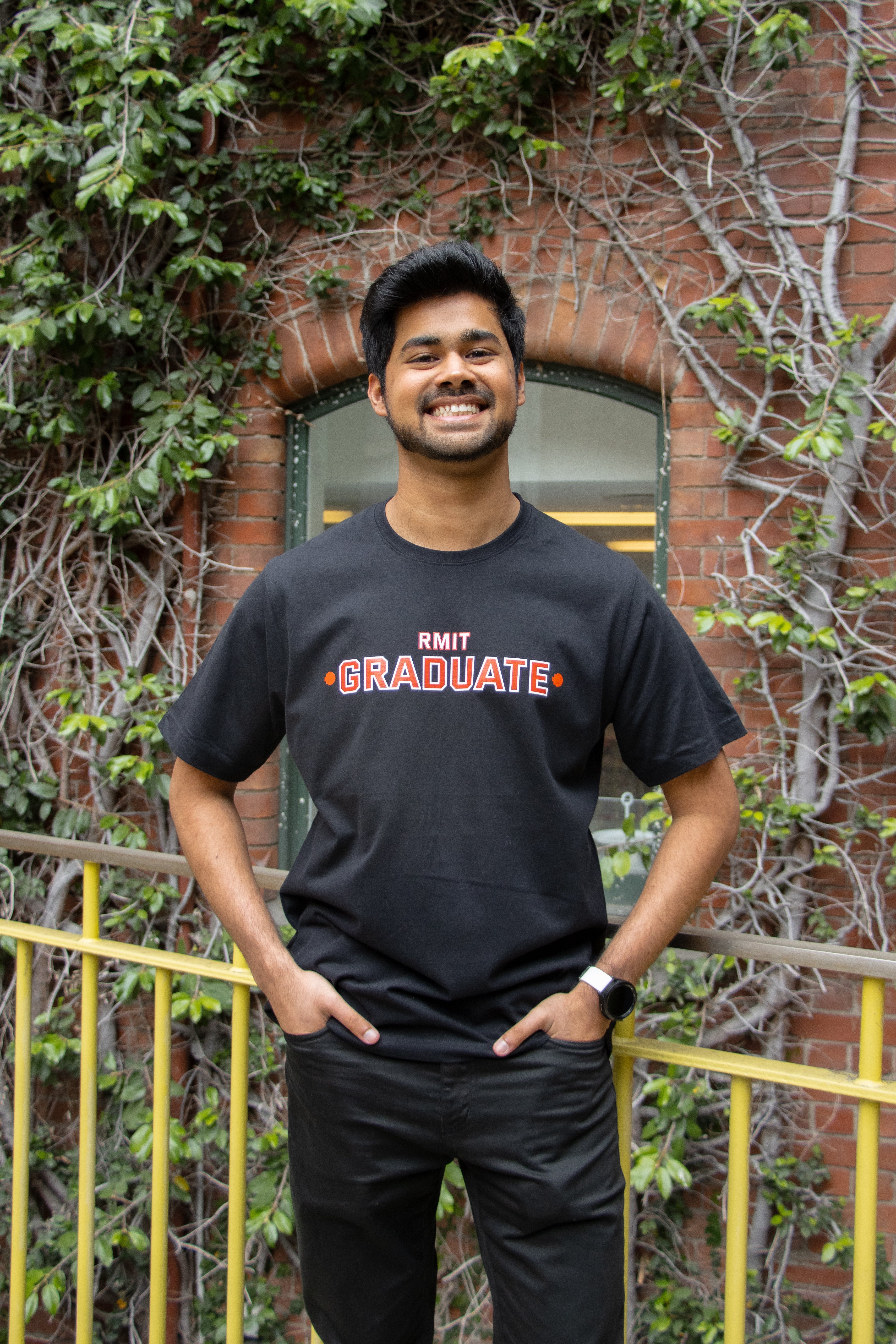 Smiling male model wearing a black RMIT Graduate T-shirt with a bold red 'RMIT Graduate' print centred on the chest. He is standing on campus in front of a brick wall with leafy vines, paired with black trousers, available at the RMIT Store.