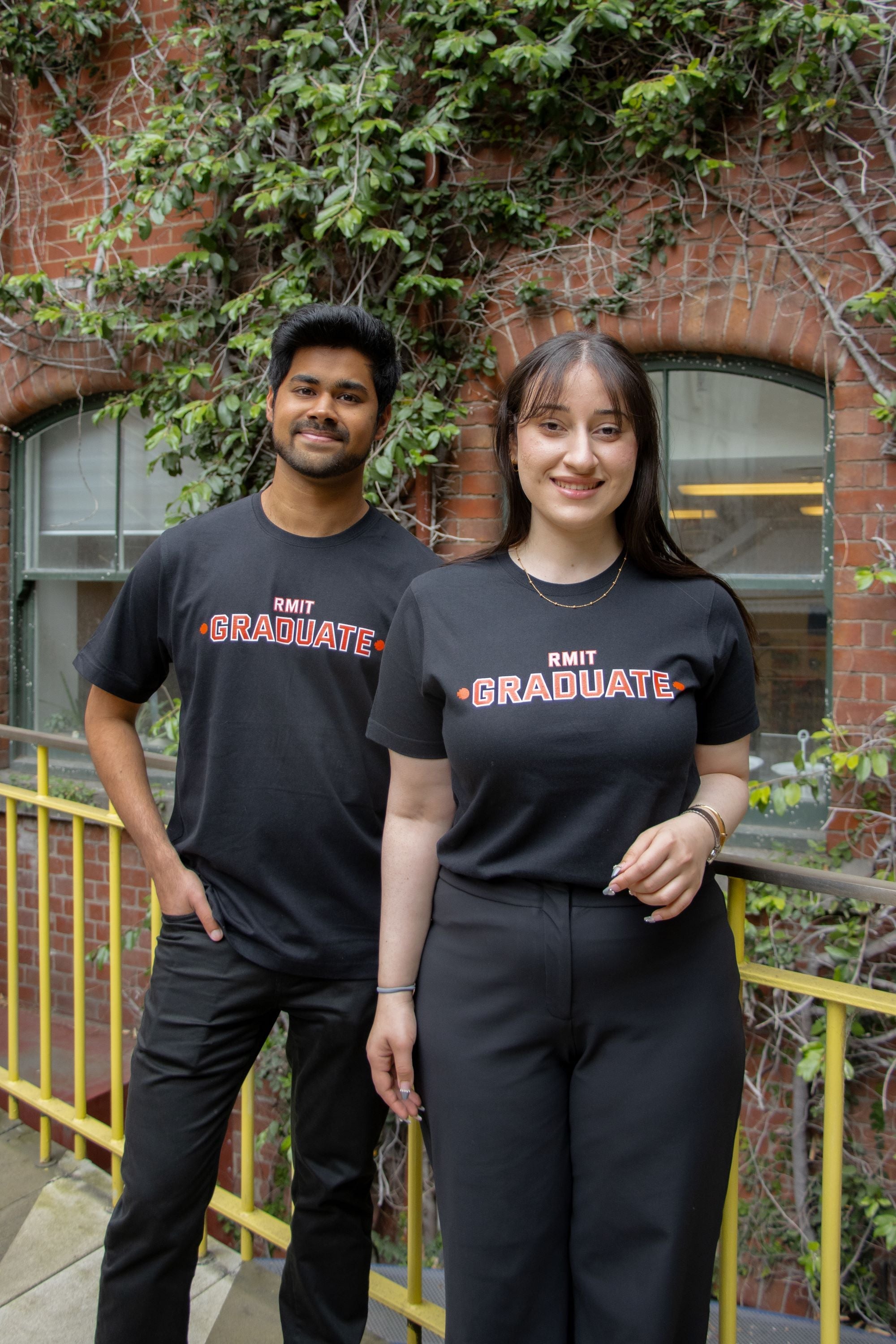Male and female models wearing matching black RMIT Graduate T-shirts on campus, each paired with black trousers. The shirts feature a bold red 'RMIT Graduate' print centred on the chest. They stand in front of a brick wall with leafy vines, available at the RMIT Store.
