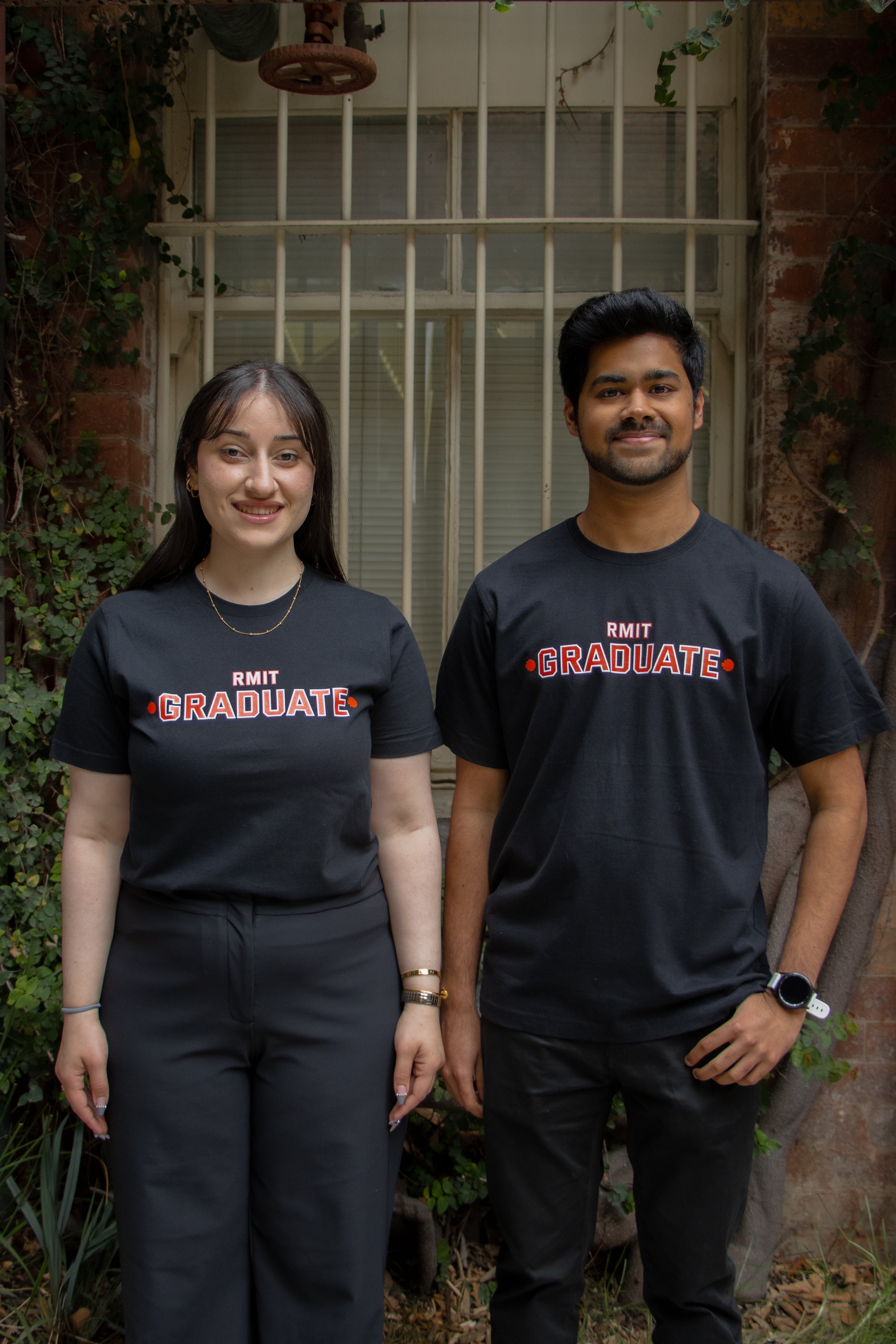 Male and female models wearing black RMIT Graduate T-shirts with red 'RMIT Graduate' print centred on the chest. They are standing side by side in front of a window set into a brick wall with leafy vines, both dressed in black trousers, available at the RMIT Store.