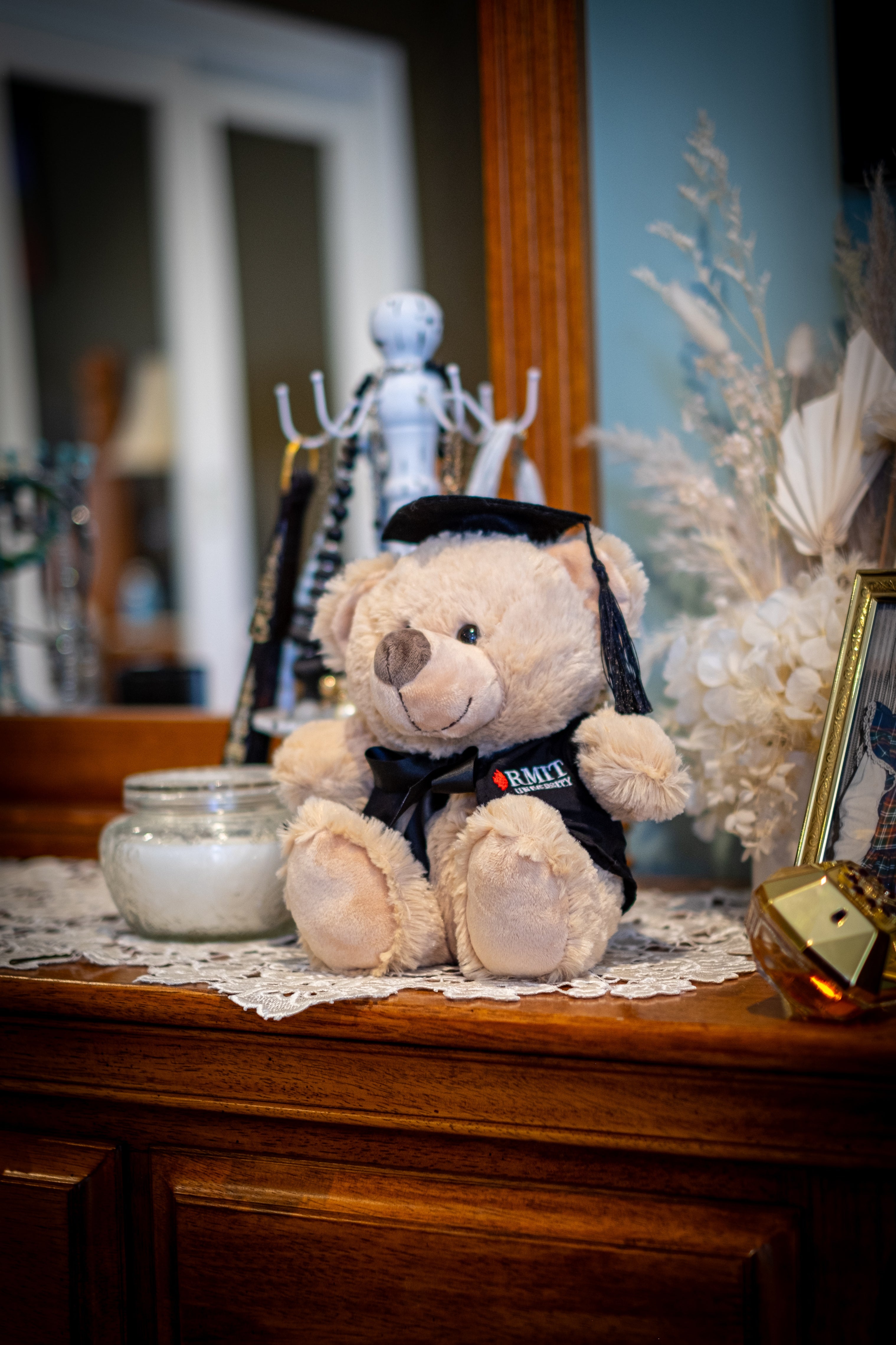 Full view of an RMIT Graduation Bear plush toy looking to its right, wearing a black graduation gown with red and white RMIT University branding on the chest and a black mortarboard with a dangling tassel, set in a warm, homey background with soft furnishings. Available at the RMIT Store.