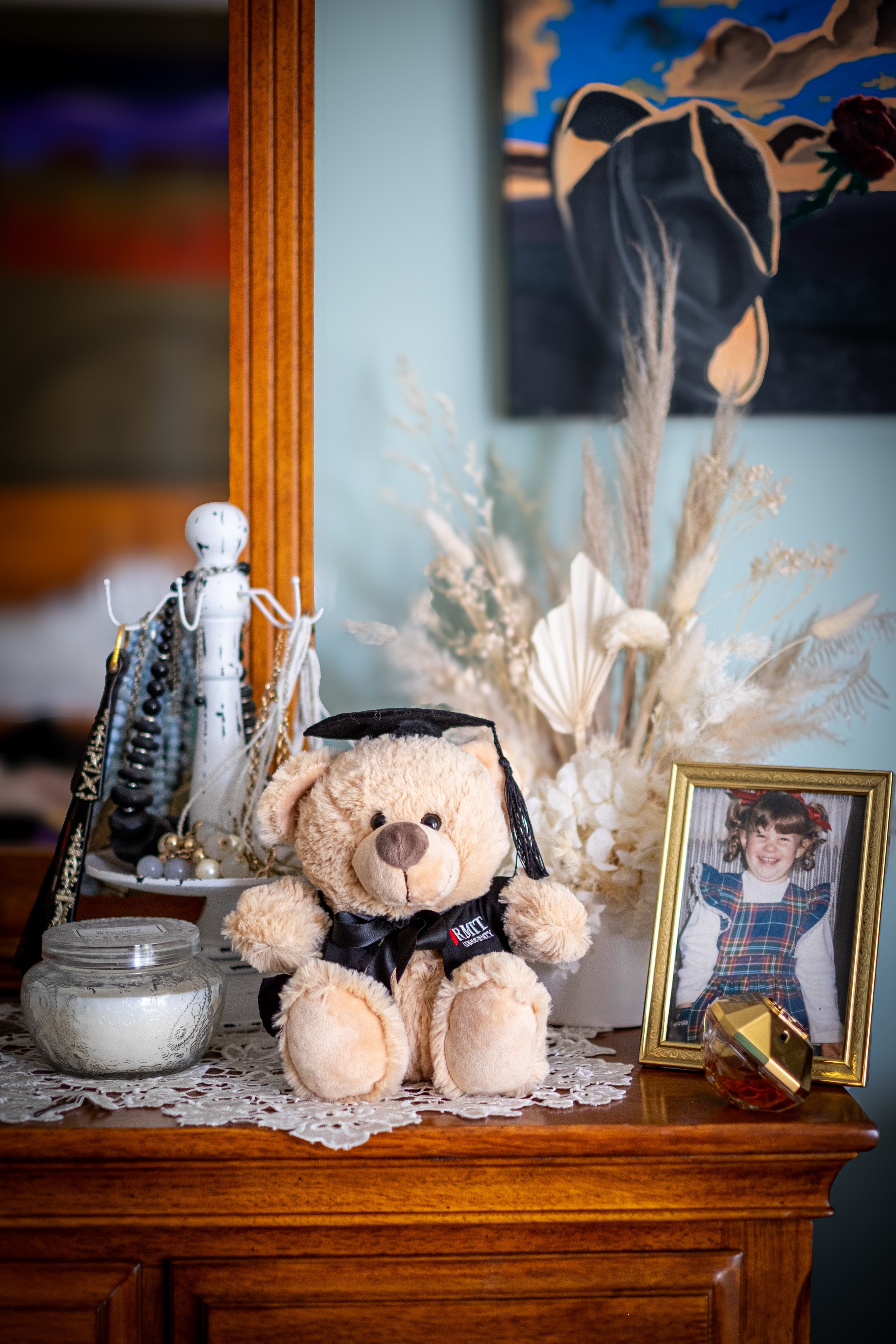 Full view of an RMIT Graduation Bear plush toy facing forward, wearing a black graduation gown with red and white RMIT University branding on the chest and a black mortarboard with a dangling tassel, positioned in a warm, homey setting with soft furnishings. Available at the RMIT Store.
