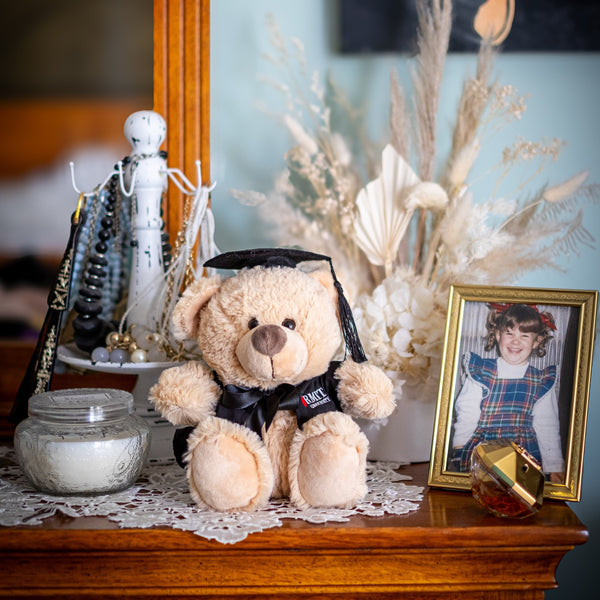 Full view of an RMIT Graduation Bear plush toy facing forward, wearing a black graduation gown with red and white RMIT University branding on the chest and a black mortarboard with a dangling tassel, positioned in a warm, homey setting with soft furnishings. Available at the RMIT Store.