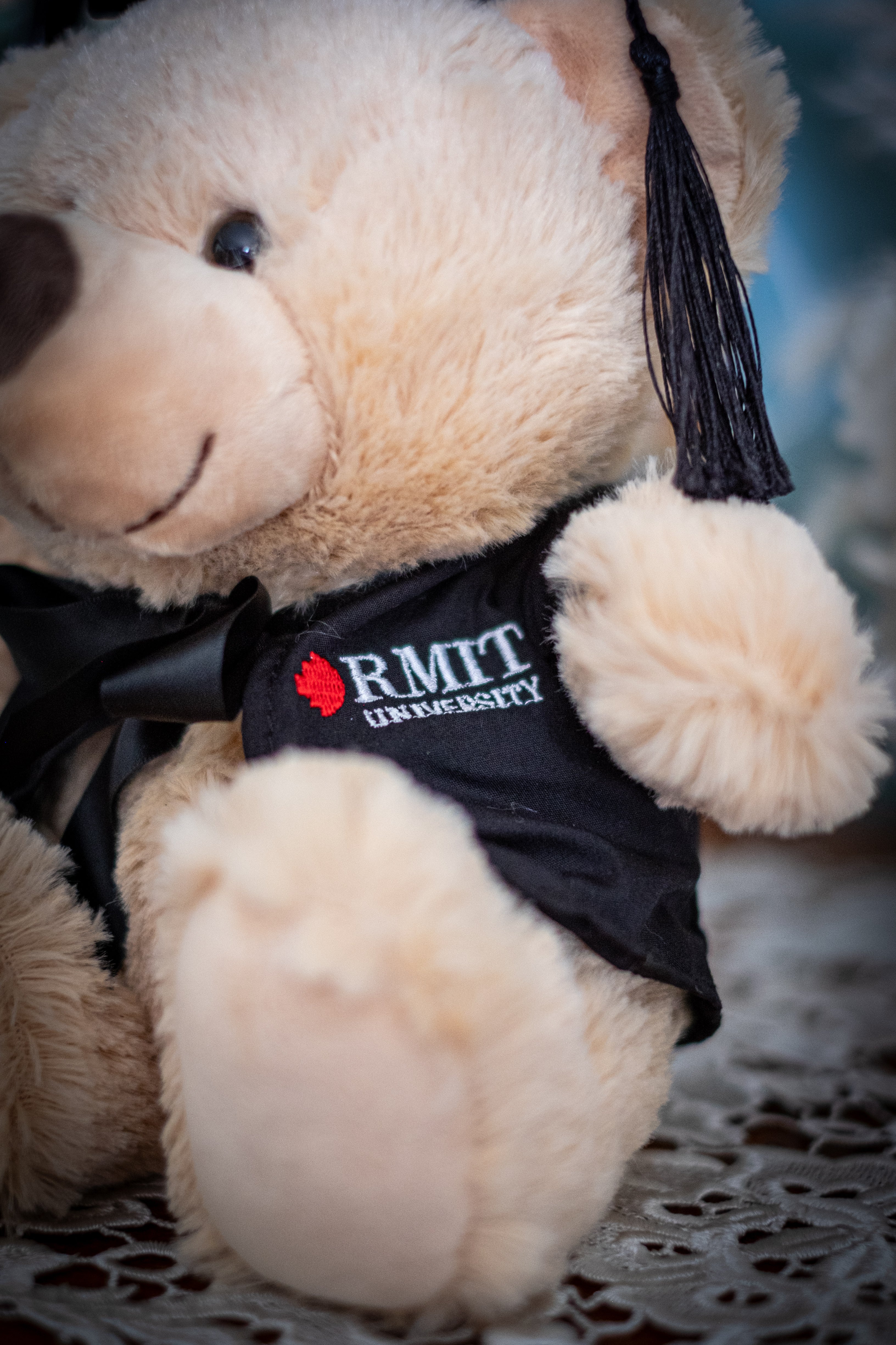 Close-up zoomed-in view of the left side of an RMIT Graduation Bear plush toy wearing a black graduation gown with red and white RMIT University branding on the chest, featuring a small dangling mortarboard tassel detail, photographed against a neutral background. Available at the RMIT Store.