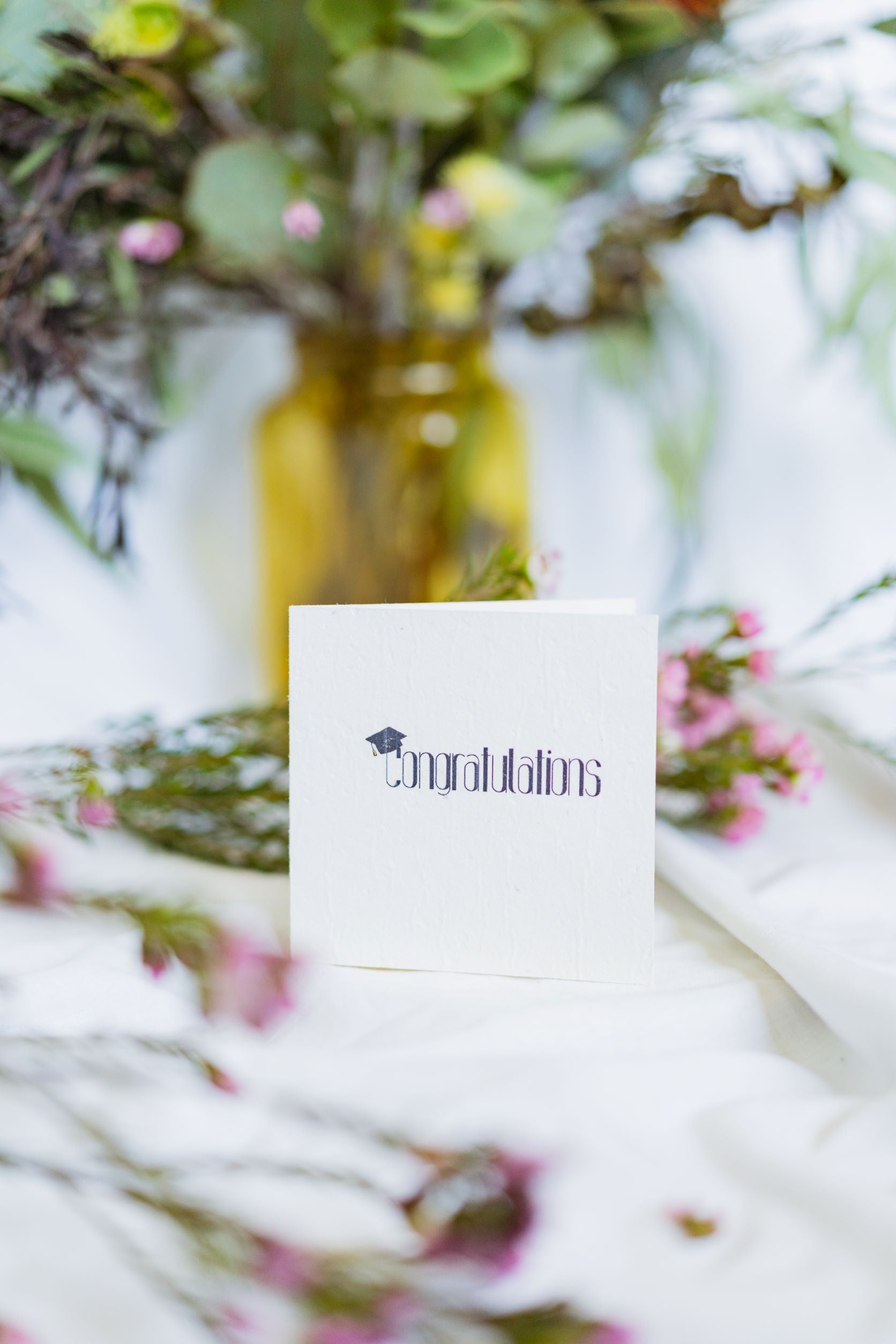 Front view of the RMIT Store Graduation Congratulations seeded card showing the word “Congratulations” in bold black centred on a white card, with a black mortarboard graduation cap perched on the letter “C,” placed against a beige fabric background with flowers next to it.