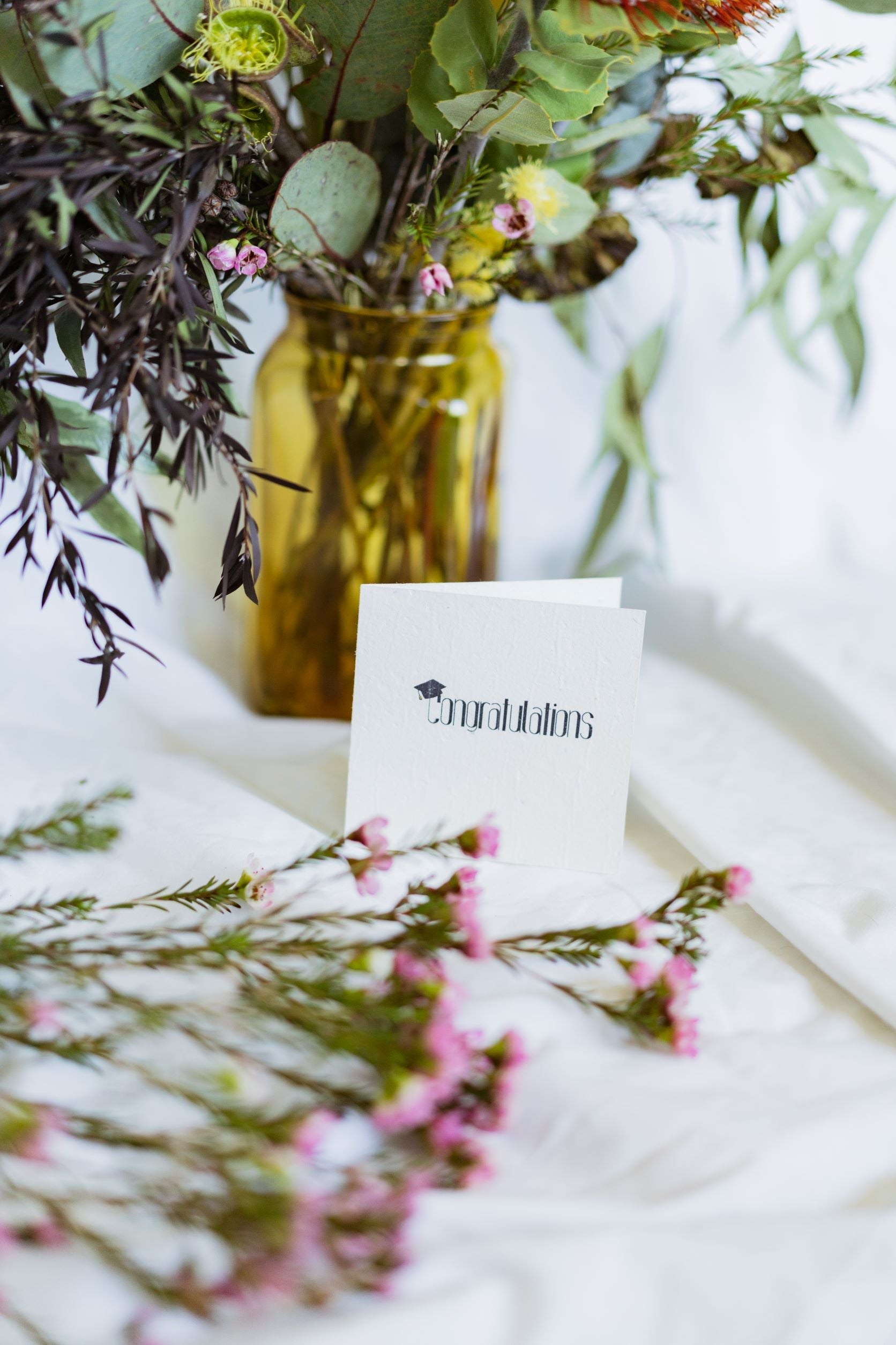 Front view of the RMIT Store Graduation Congratulations seeded card showing the word “Congratulations” in bold black centred on a white card, with a black mortarboard graduation cap perched on the letter “C,” placed against a beige fabric background with flowers next to it.