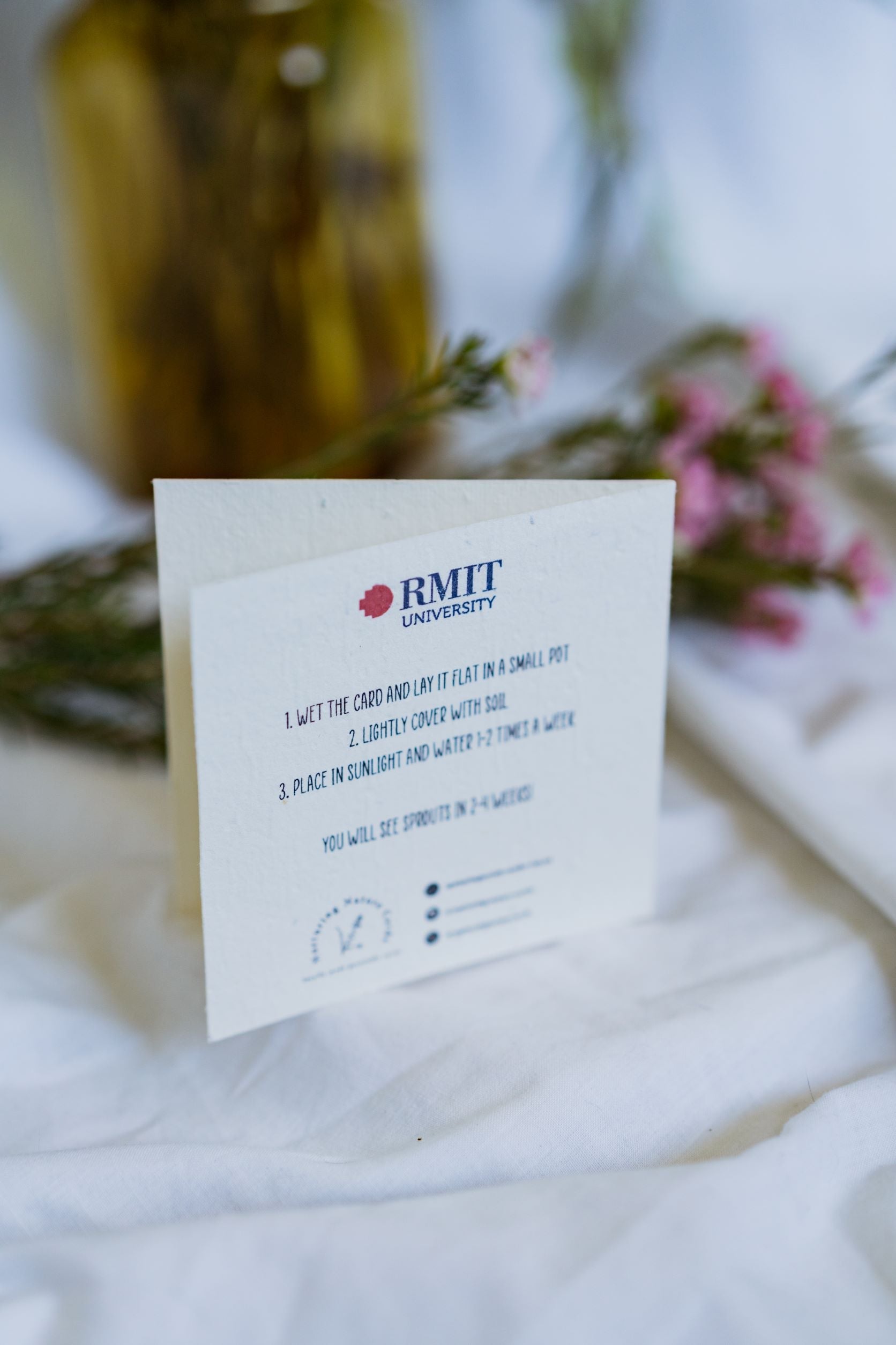 Rear view of the RMIT Store Graduation Congratulations Card in seeded paper, displayed upright against a light beige fabric background. The RMIT University Logo is on the top of the card.