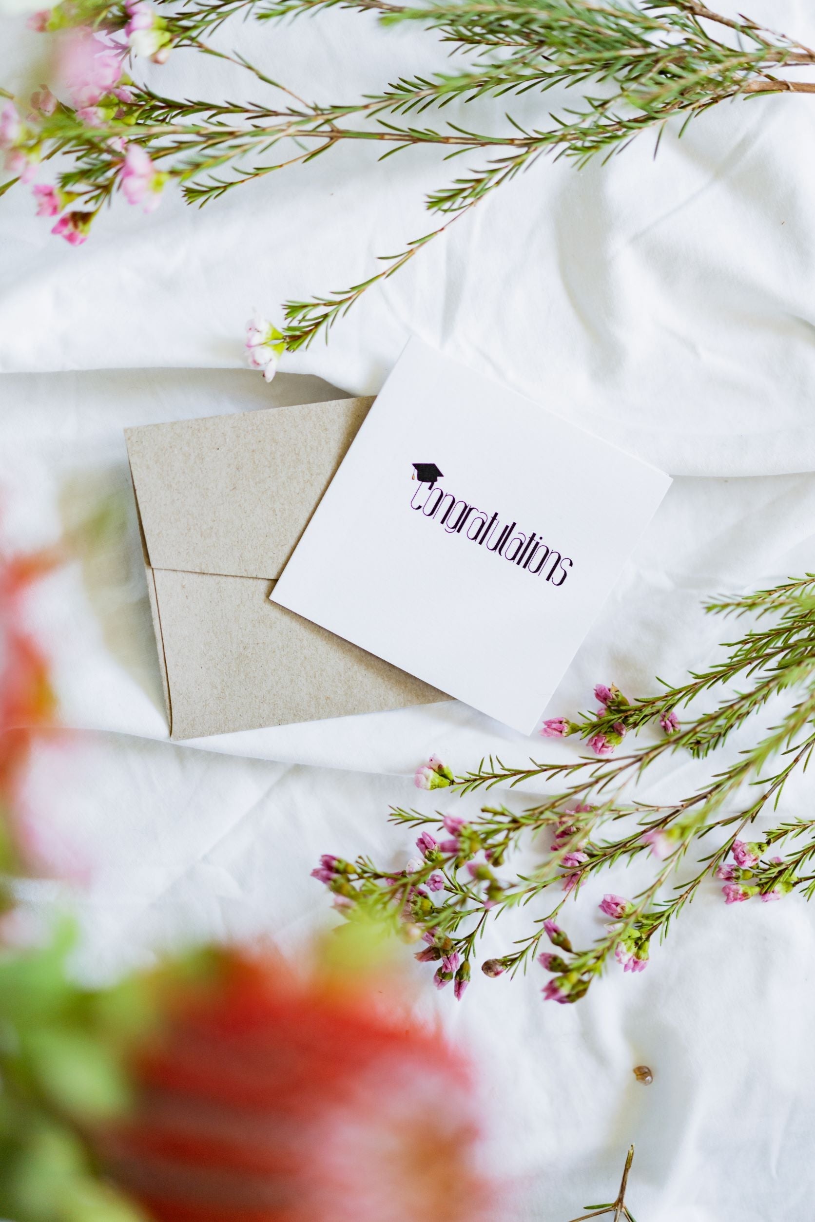 Front view of the RMIT Store Graduation Congratulations card, unseeded version, resting on a light brown envelope against a white fabric background, featuring the word “Congratulations” in black with a mortarboard graduation cap incorporated into the letter “C”.