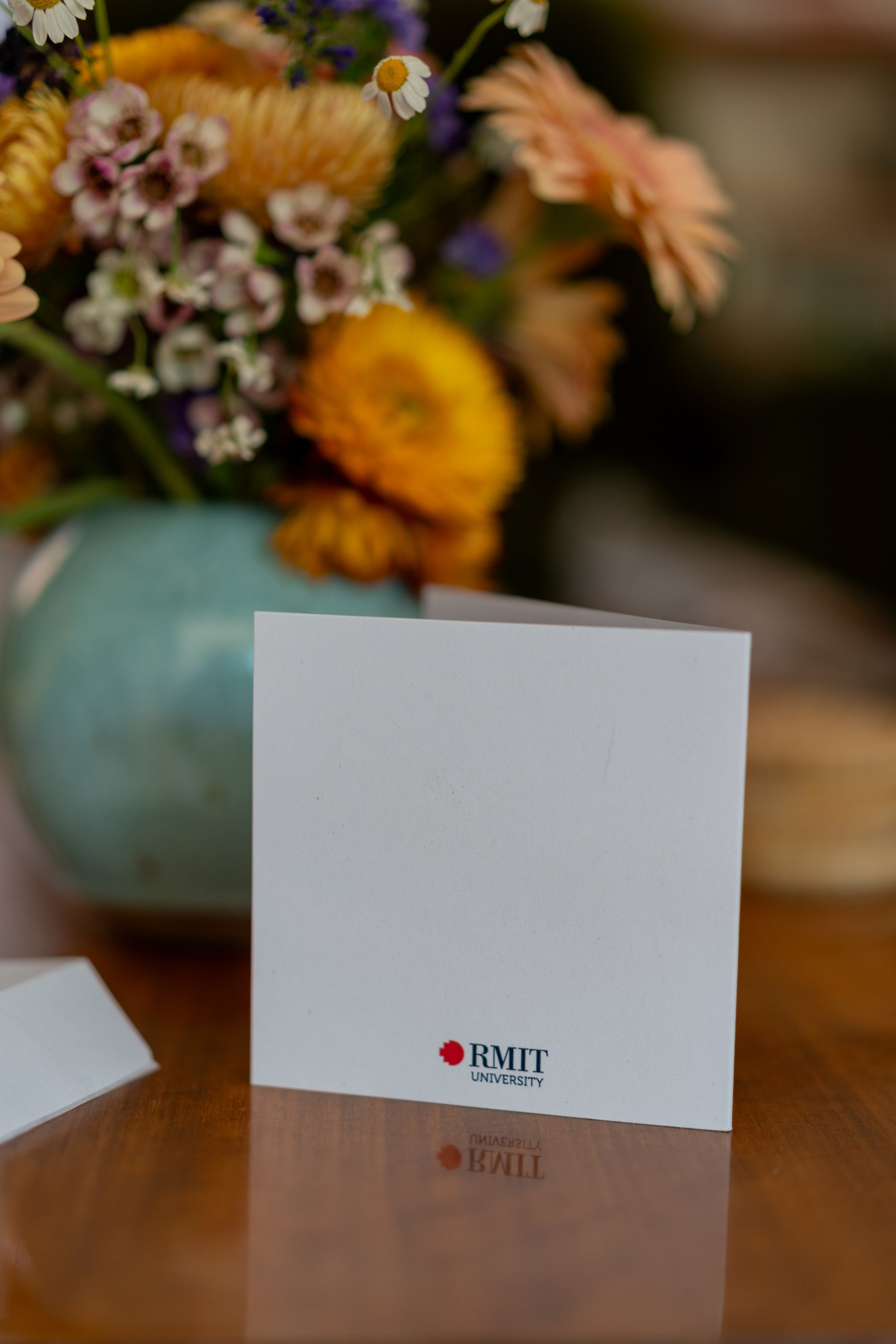 Back view of the RMIT Store graduation parade card standing upright on a wooden tabletop. The card has a plain white surface with the RMIT logo printed at the bottom centre. A vase with flowers is partially visible to the right, adding a soft decorative touch to the setting.