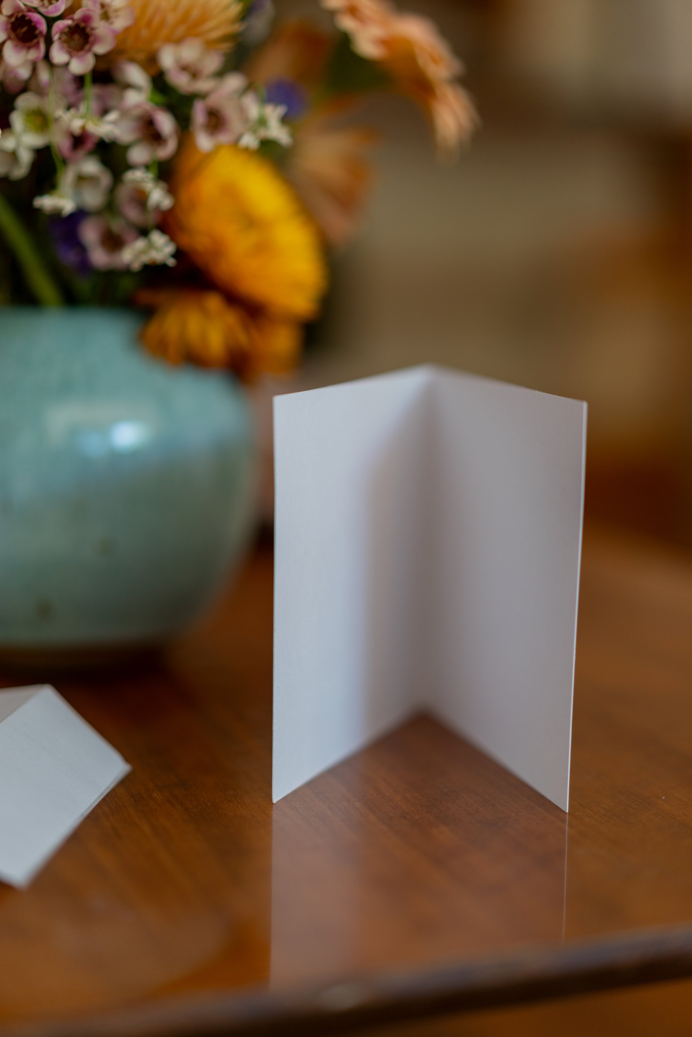 Graduation parade card from the RMIT Store standing upright on a wooden tabletop, with a vase containing flowers positioned to its right. The card is open to reveal a plain white interior. The warm setting highlights the card’s simple and elegant design.