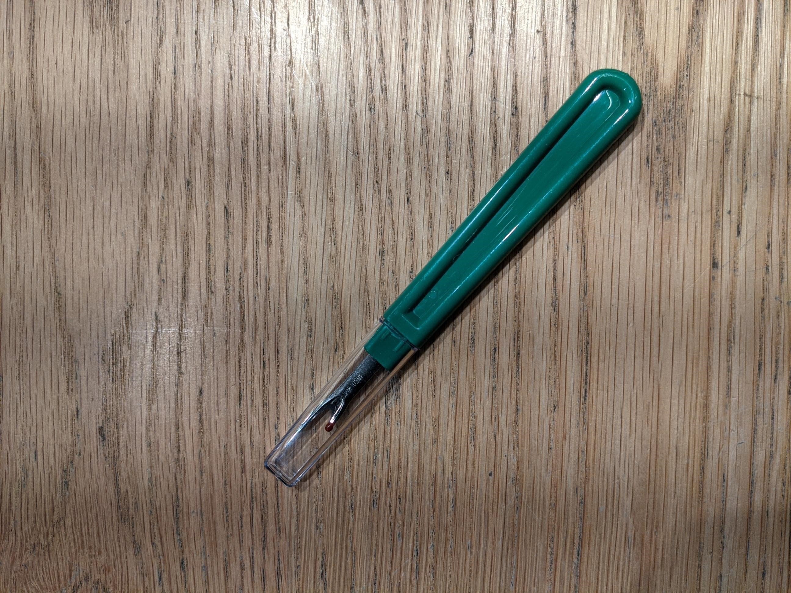 Green seam unpicker with a curved plastic handle and protective clear cap, displayed diagonally on a beige fabric tabletop. The metal blade features a red safety ball on the shorter tip. Sold through the RMIT Store.