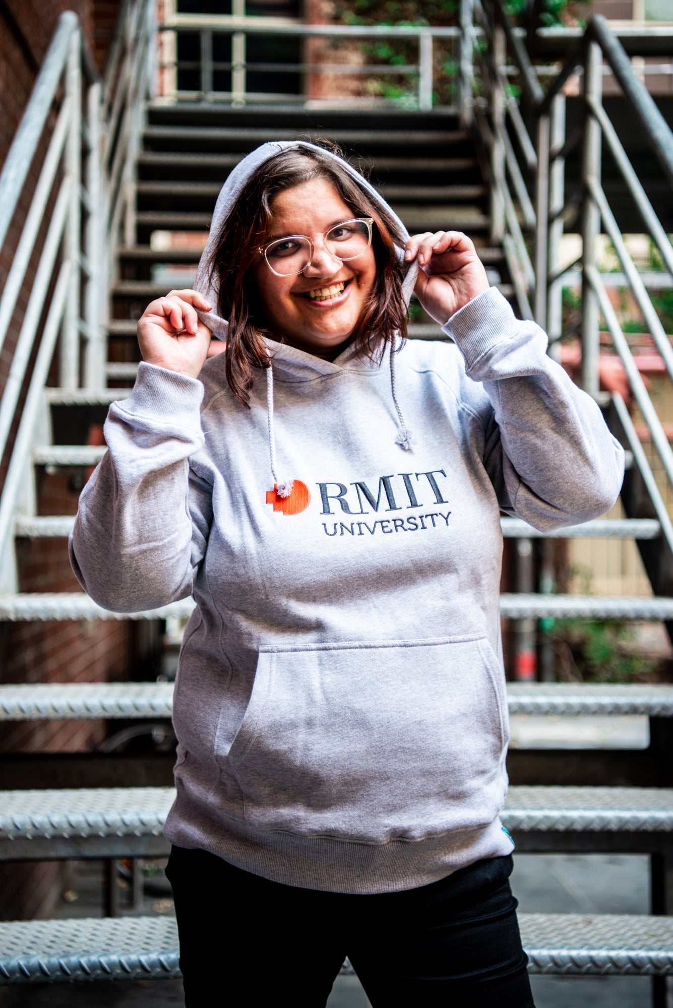 Female model wearing a grey Fairtrade hoodie from the RMIT Store, facing forward with both hands holding the edges of the hood. She is smiling gently, and the hoodie features a relaxed fit with a red and white embroidered RMIT University logo centred on the chest. 