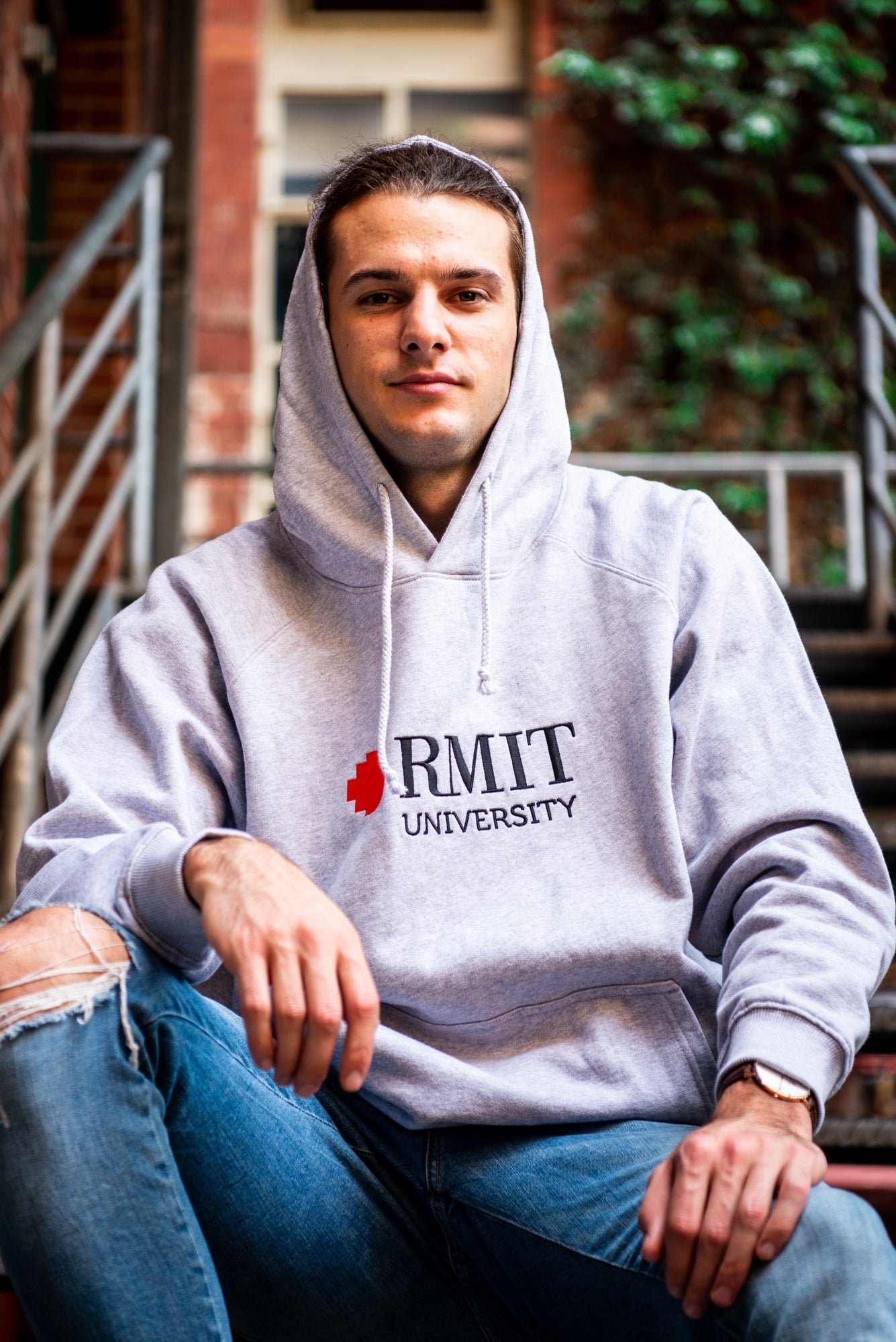 Male model wearing a grey Fairtrade hoodie from the RMIT Store, facing forward with a relaxed expression with the hood up. The hoodie features a red and white embroidered RMIT University logo centred on the chest. 