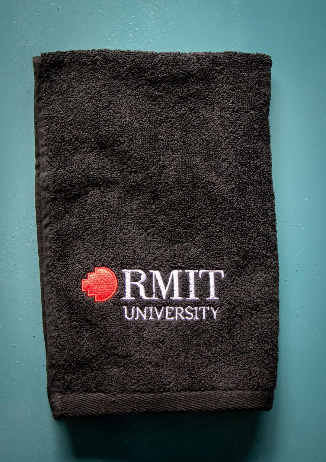 Black RMIT University hand towel with embroidered logo from the RMIT Store, displayed on a teal background, highlighting its soft texture and elegant branding.