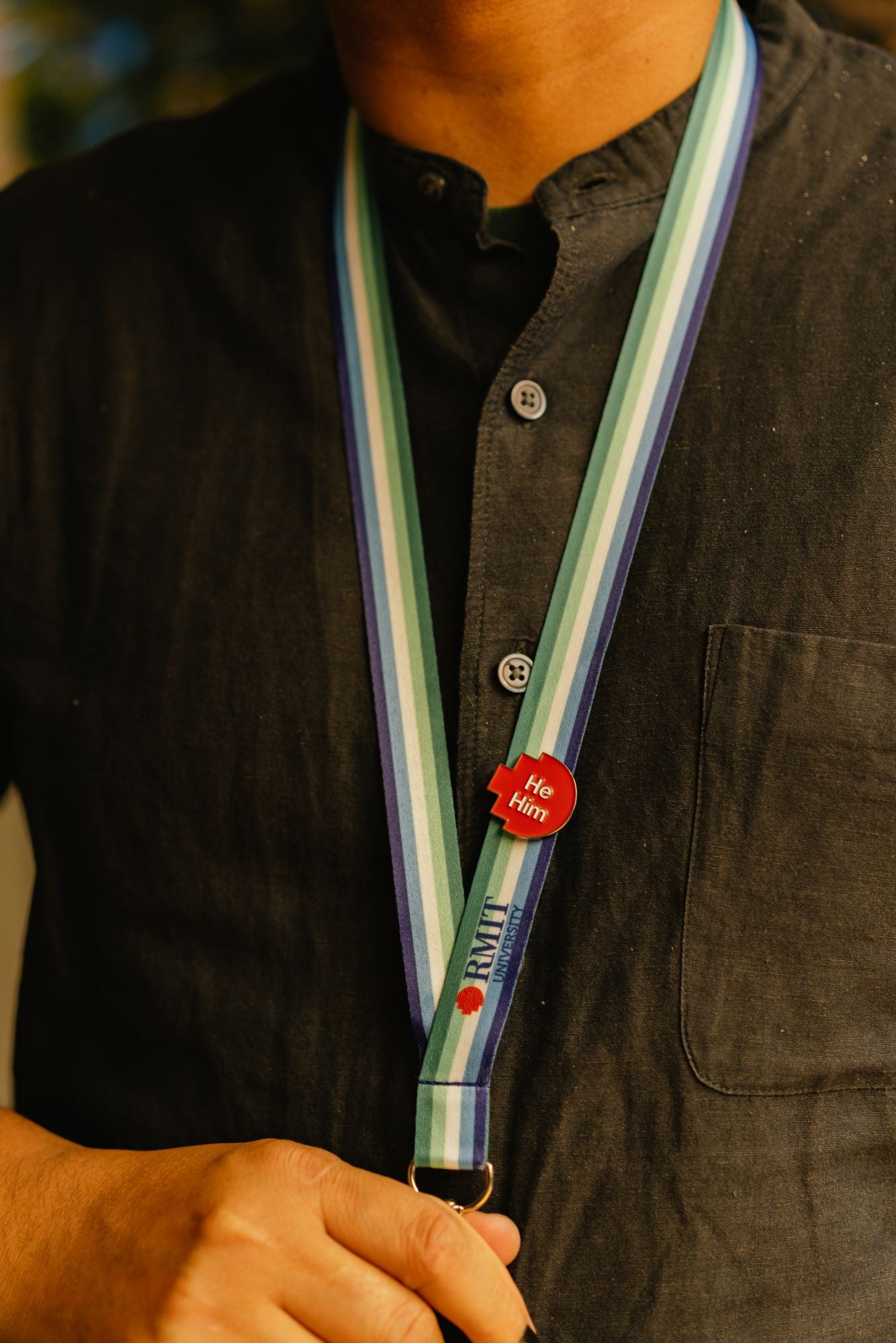 Close-up of a model wearing a red pixelated “he/him” pronoun pin from the RMIT Store, attached to a Trans-Inclusive Gay Men’s Pride lanyard featuring blue, white, and green horizontal stripes. The model’s left hand is holding the end of the lanyard.