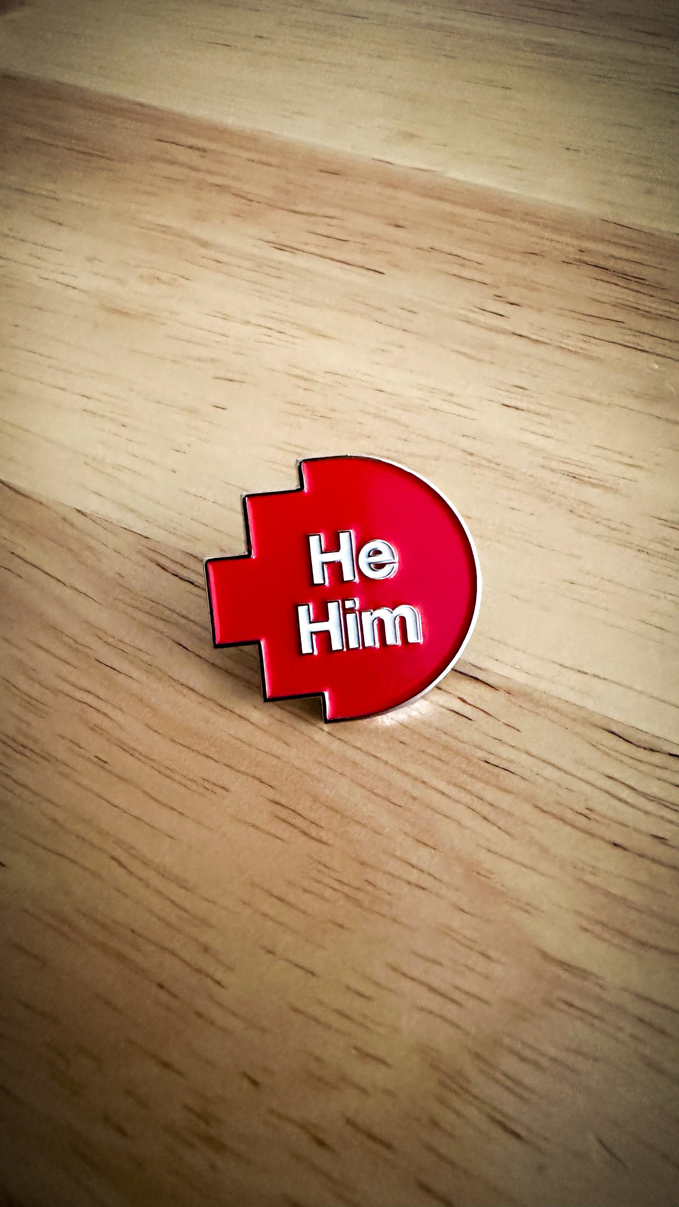Close-up of a he/him pronoun pin from the RMIT Store, featuring white printed text “He/Him” on a red pixelated background. The pin is made of metal with a smooth finish and is displayed on a wooden surface background.