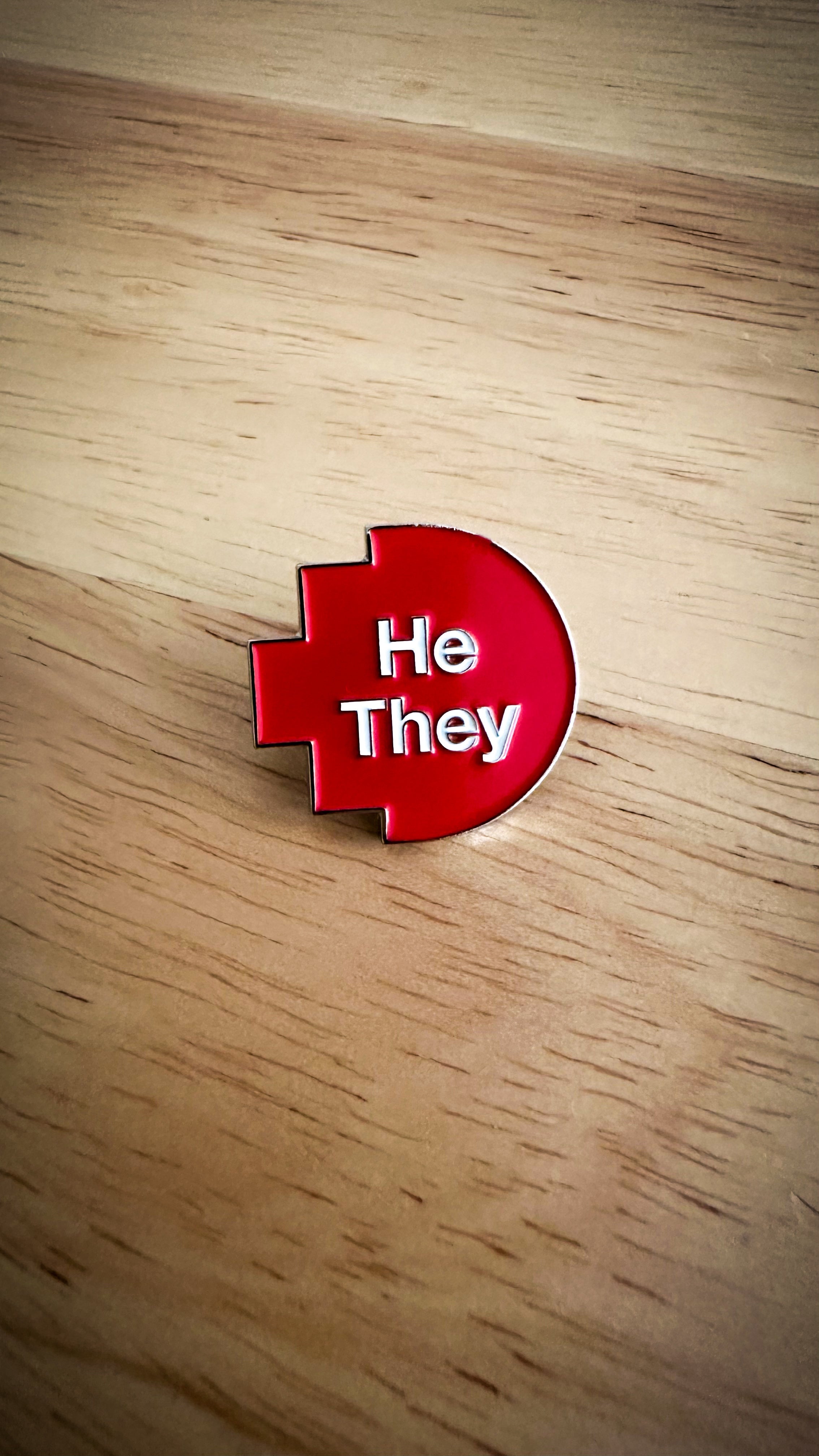 Close-up of a He/They pronoun pin from the RMIT Store, featuring white printed text “He/They” on a red pixelated background. The pin is made of metal with a smooth finish and is displayed on a wooden surface background.