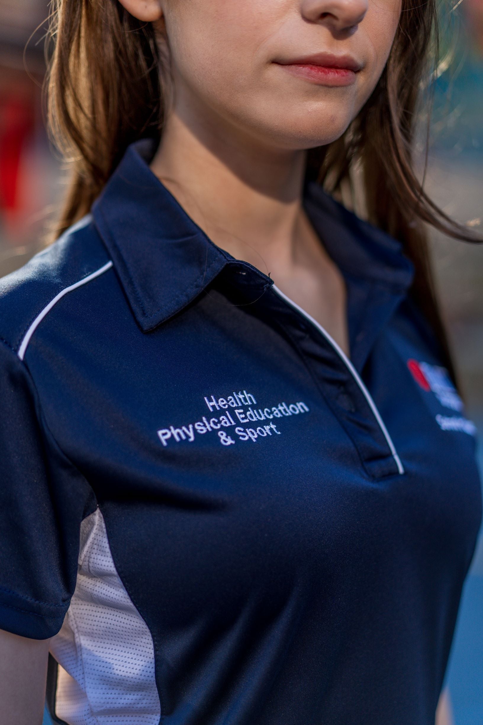 Close-up view of the female model's right chest area on the RMIT Store Health, Physical Education & Sport Uniform Polo. The navy polyester polo features detailed white embroidery that reads “Health, Physical Education & Sport.” The texture of the stitching is visible, with part of the collar and sleeve also in frame.