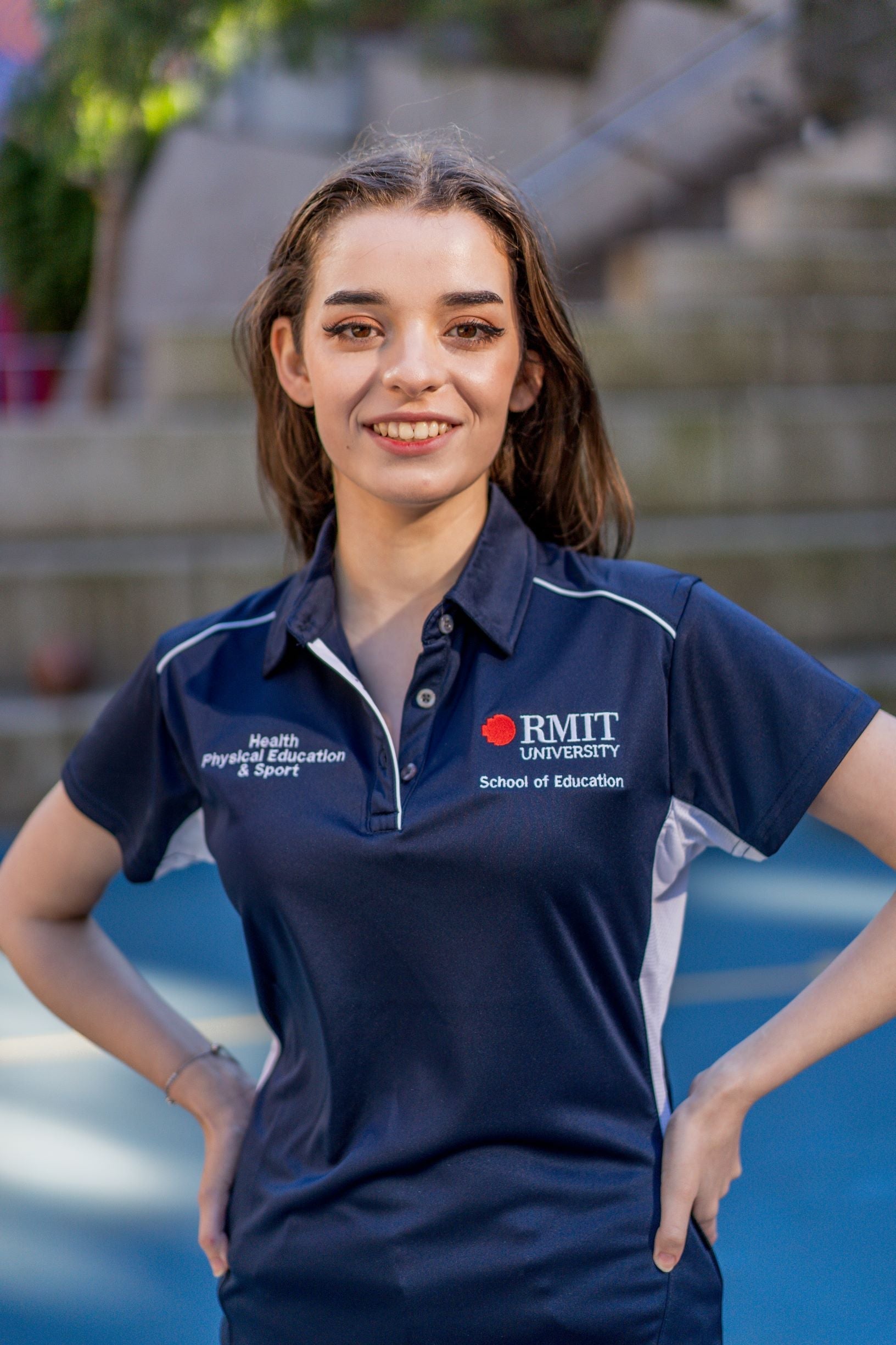 Front view of a female model wearing the navy polyester Health, Physical Education & Sport Uniform Polo from the RMIT Store, featuring the white and red RMIT University logo on the left chest and white embroidered “Health, Physical Education & Sport” text on the right chest. The model is standing outdoors on campus with hair down.