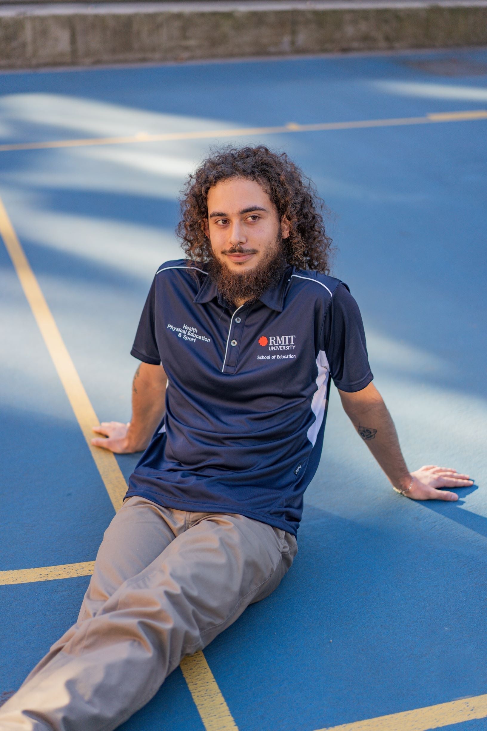 Male model sitting on the ground on campus wearing the navy Health, Physical Education & Sport Polo from the RMIT Store paired with beige pants, showing a full front view of the polo.
