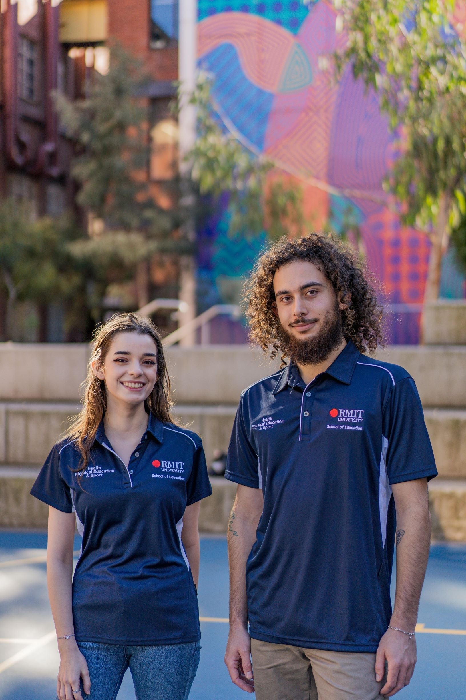 Male and female model on campus wearing the RMIT Store Health, Physical Education & Sport Uniform Polo. The navy polyester polo features the RMIT University logo in red and white on the left chest, with “Health, Physical Education & Sport” printed in white on the right chest. Both models are outdoors in a university setting.