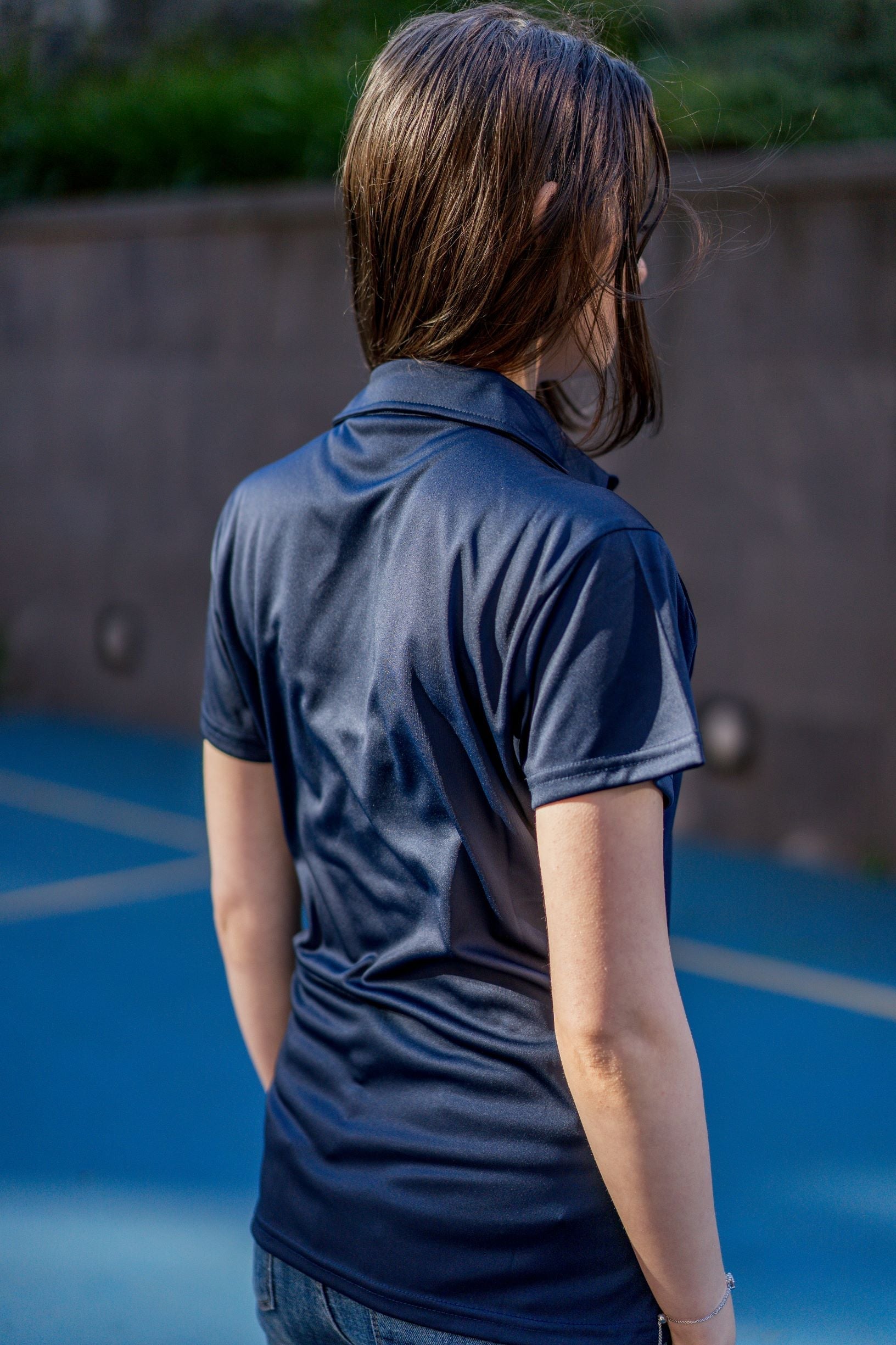 Side-back view of a female model standing in the sun on campus, wearing the plain navy RMIT Store Health, Physical Education & Sport Uniform Polo. The polyester polo has no visible logos or text on this side, and the model’s hair is worn down.