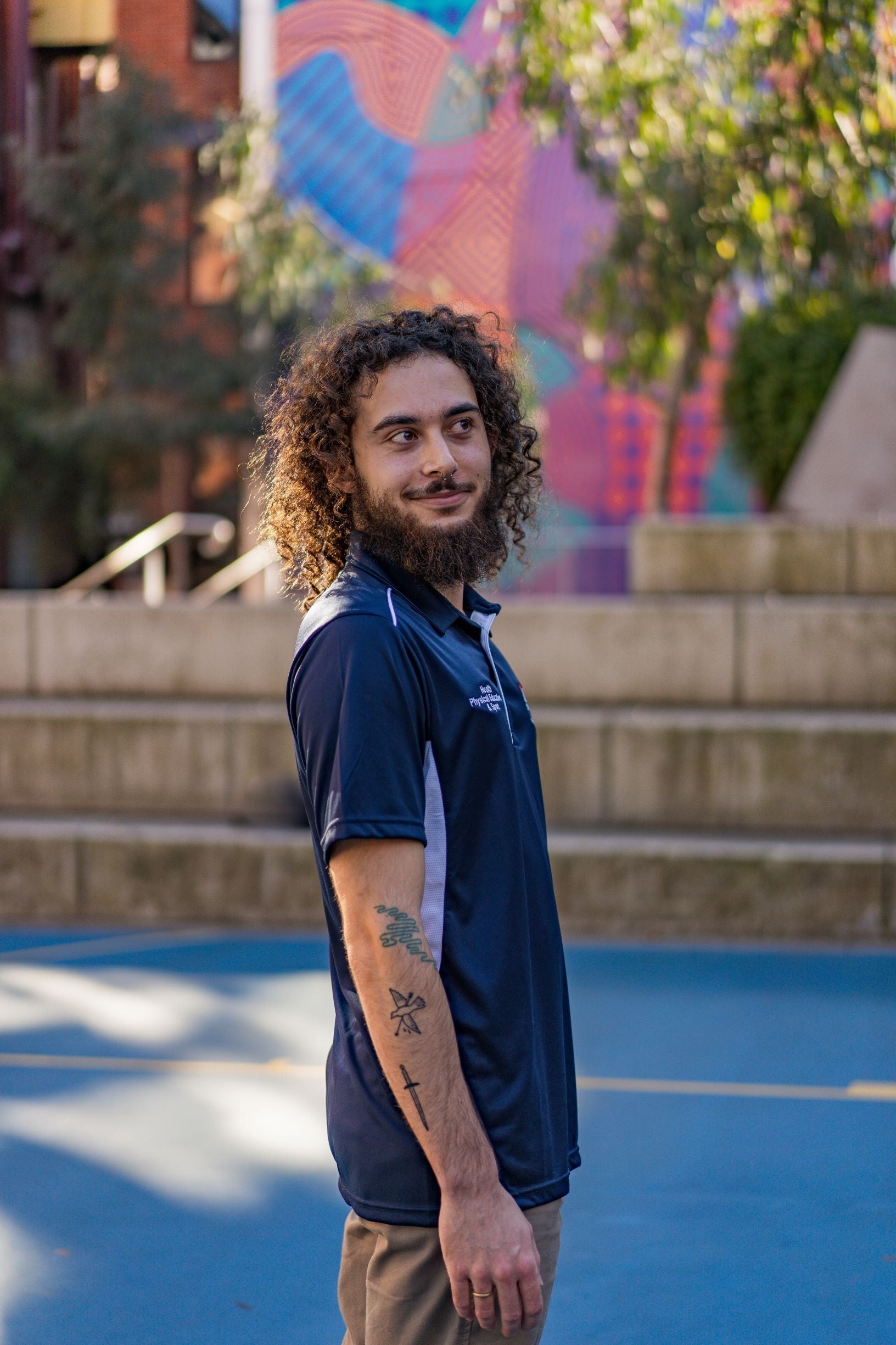 Side view of a male model wearing the navy Health, Physical Education & Sport Polo from the RMIT Store, looking to his left. The image shows the short sleeve and side profile of the polo with visible "Health, Physical Education & Sport" on the right chest.
