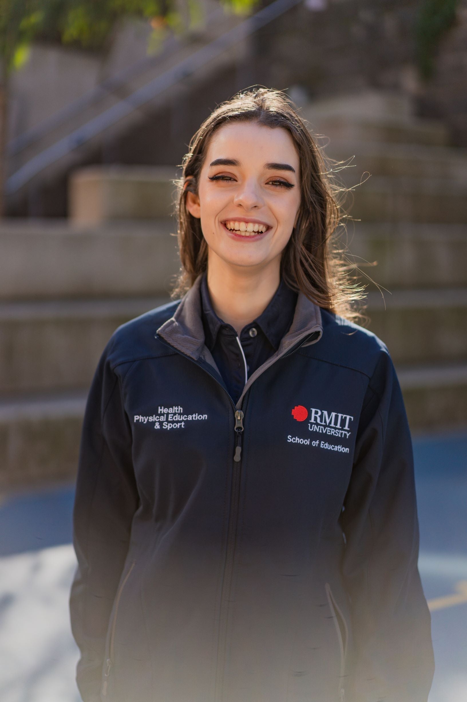 Front view of a female model smiling and wearing the navy Health, Physical Education & Sport Uniform – Jacket with RMIT University logo and “School of Education” embroidered on the left chest, and “Health, Physical Education & Sport” embroidered on the right chest, outdoors on campus. Available at the RMIT Store.