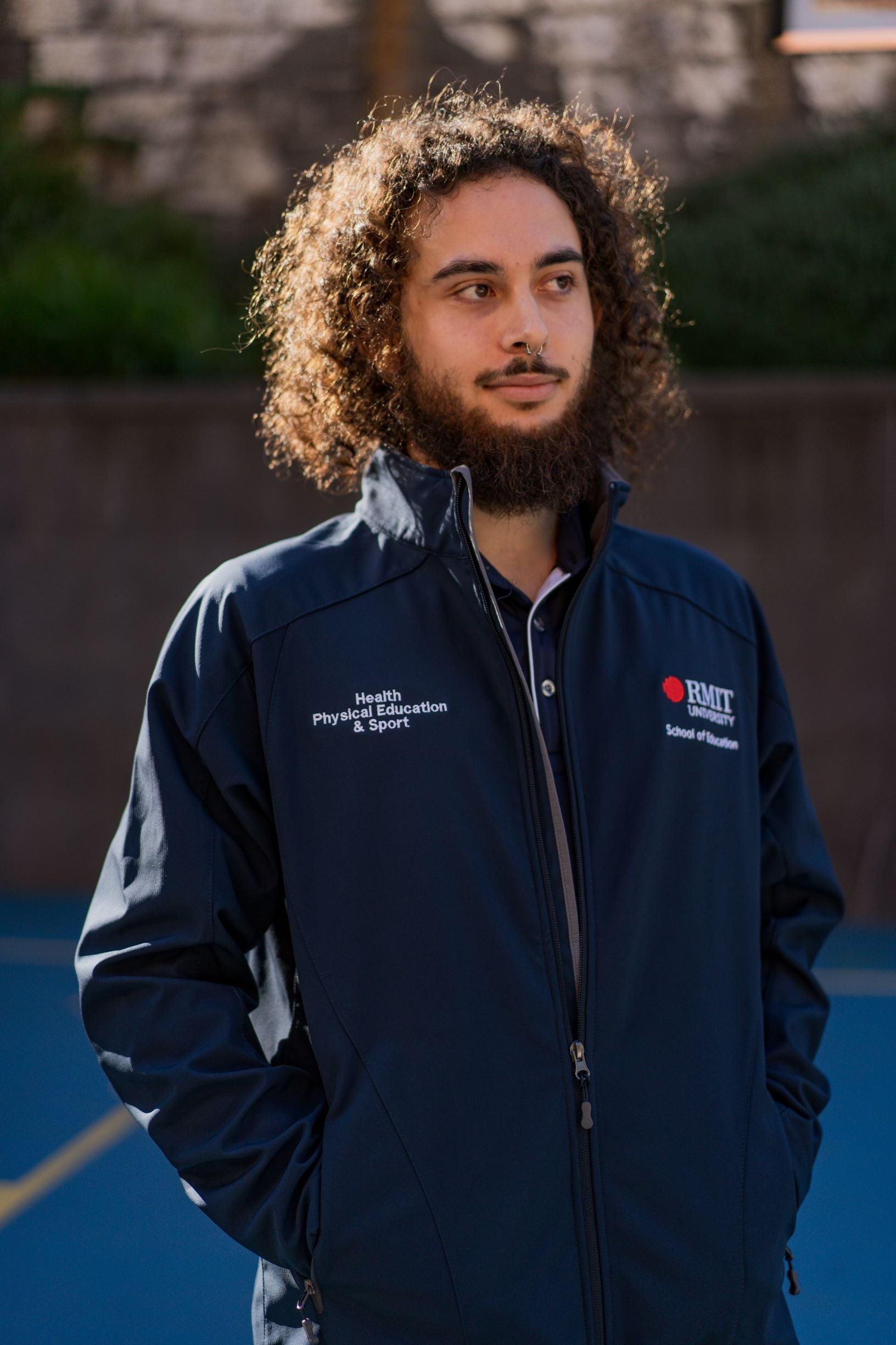 Side-turned front view of a male model wearing the navy Health, Physical Education & Sport Uniform – Jacket, looking to his left. The left chest shows the RMIT University logo with “School of Education” embroidered beneath, and the right chest displays “Health, Physical Education & Sport”. Photographed outdoors in a campus setting. Available at the RMIT Store.