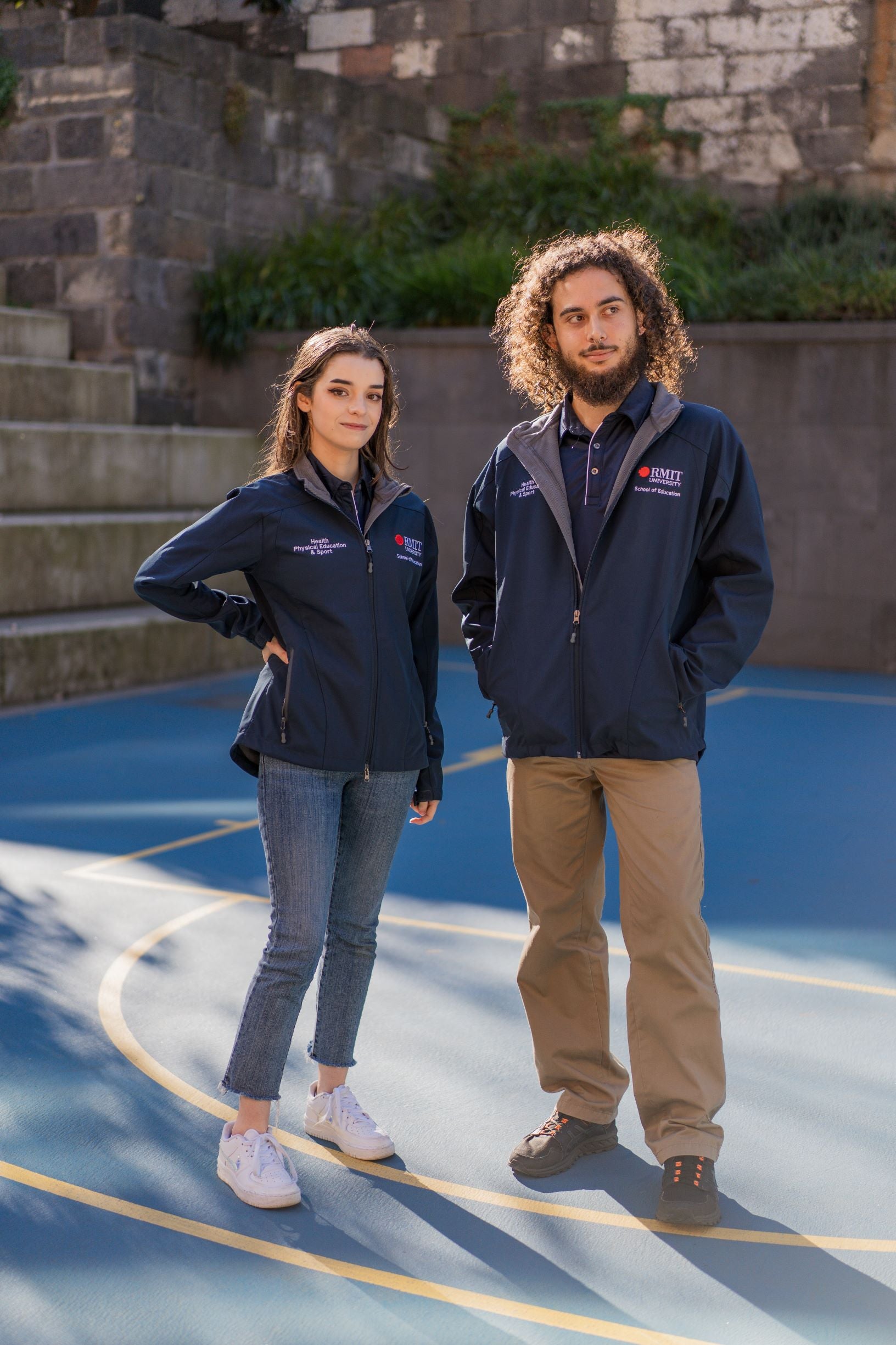 Full front view of a female model with one hand on her waist looking into the camera and a male model looking to his left, both wearing the navy Health, Physical Education & Sport Uniform – Jacket. The left chest features the RMIT University logo with “School of Education” embroidered beneath, and the right chest reads “Health, Physical Education & Sport”. Captured outdoors on campus. Available at the RMIT Store.