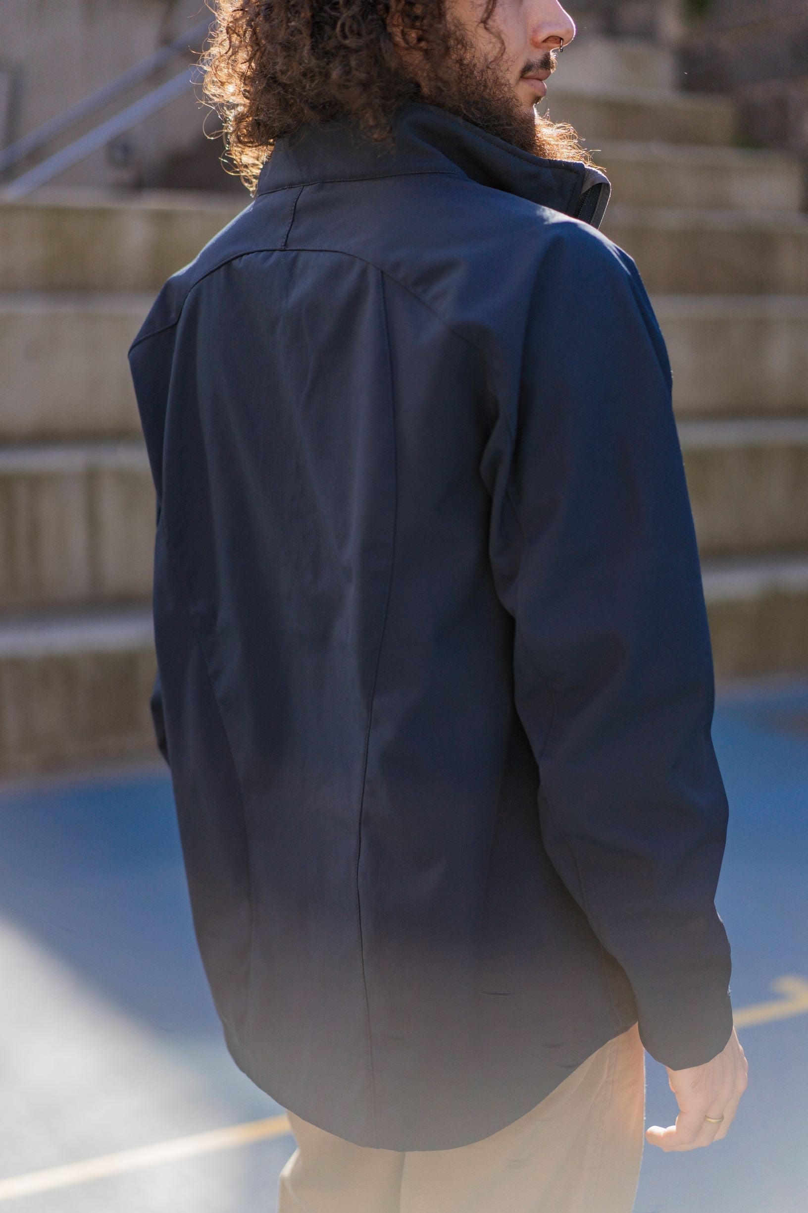 Rear view of a male model wearing the navy Health, Physical Education & Sport Uniform – Jacket, standing outdoors on campus with his body slightly turned to the right. The jacket features a clean, plain back. Model wears beige trousers. Available at the RMIT Store.