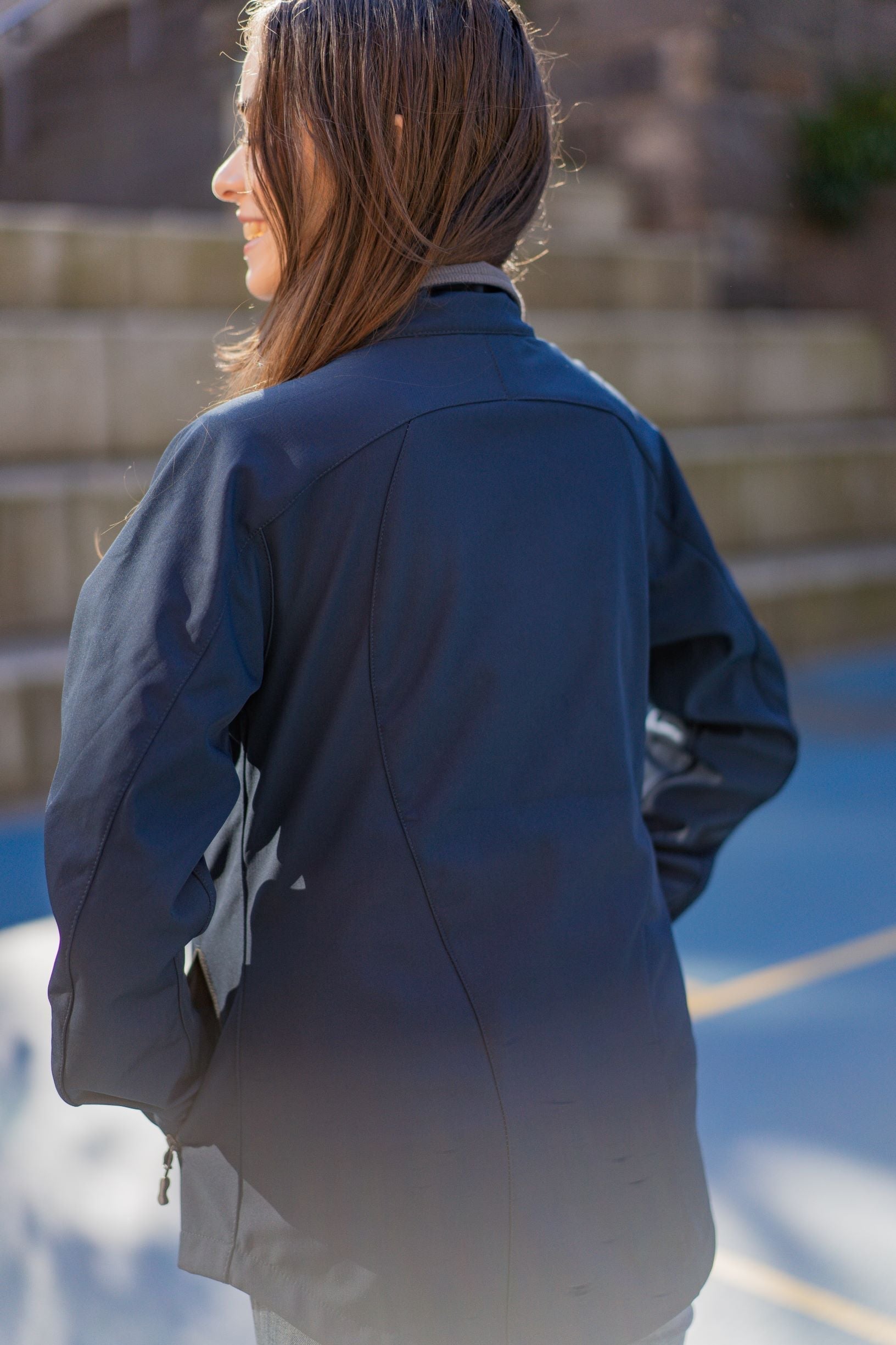 Rear view of a female model wearing the navy Health, Physical Education & Sport Uniform – Jacket, slightly turned to her left outdoors on campus. Available at the RMIT Store.