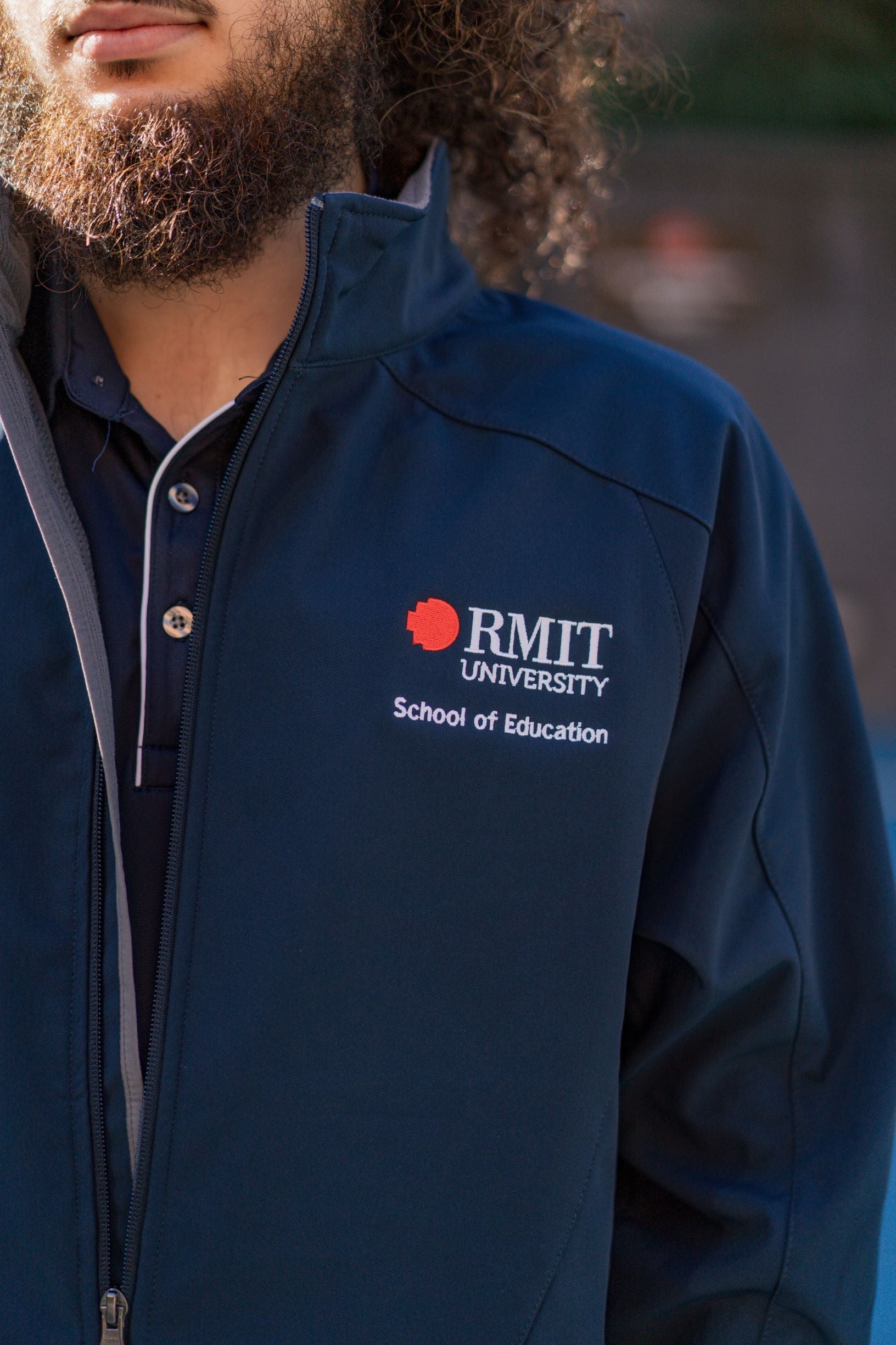 Zoomed-in view of the left chest on the navy Health, Physical Education & Sport Uniform – Jacket, featuring white embroidered "School of Education" text positioned beneath the white and red RMIT University logo embroidery. Fabric texture is smooth and matte. Available at the RMIT Store.