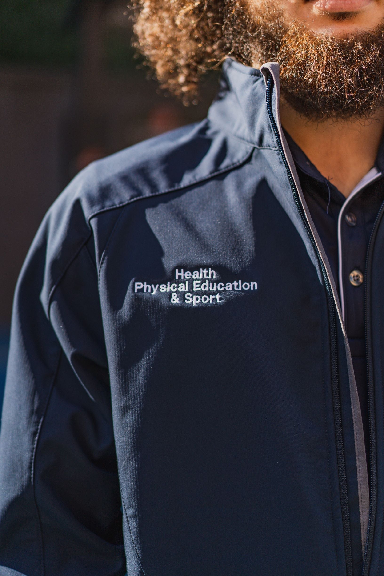 Zoomed-in view of the right chest on the navy Health, Physical Education & Sport Uniform – Jacket, showing white embroidered text reading "Health, Physical Education & Sport". Fabric texture is smooth and matte. Available at the RMIT Store.