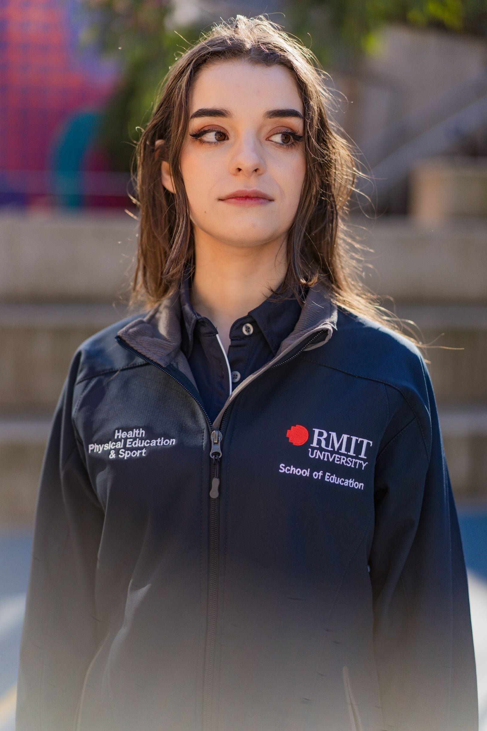 Close-up front view of a female model wearing the navy Health, Physical Education & Sport Uniform – Jacket, looking to her left. The left chest shows embroidered RMIT University logo with “School of Education” underneath, and the right chest features embroidered “Health, Physical Education & Sport.” Outdoor campus background. Available at the RMIT Store.