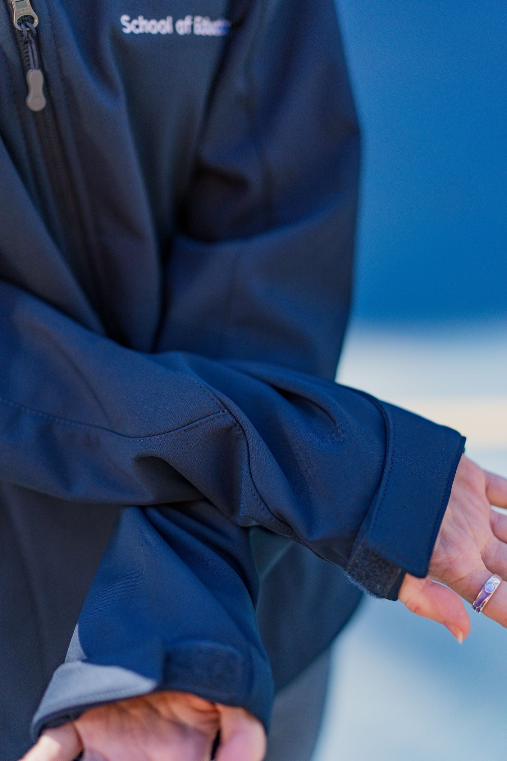 Close-up view of the navy Health, Physical Education & Sport Uniform – Jacket sleeves worn by a model, showing adjustable cuffs and subtle fabric texture. The model’s right hand is visible wearing a silver ring. Background is an outdoor campus setting. Available at the RMIT Store.