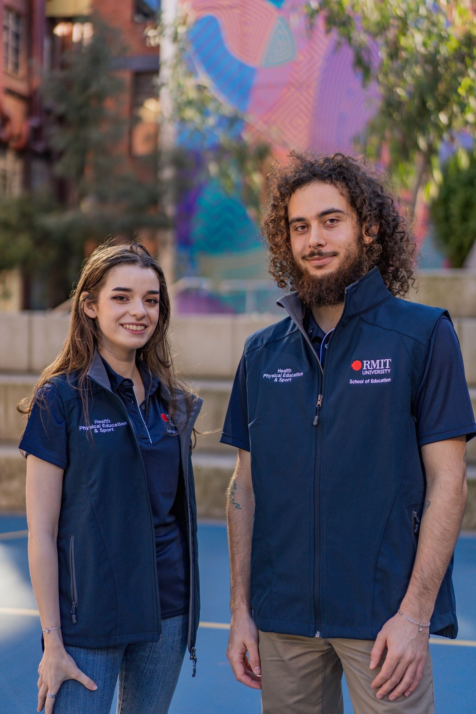 Front view of a male and a female model wearing the Health, Physical Education & Sport Uniform – Vest, both looking into the camera. The female model has her head slightly turned to the left, and the male model slightly to the right. Both navy vests feature white embroidered “Health, Physical Education & Sport” on the right chest and the RMIT University logo with “School of Education” on the left chest. Available at the RMIT Store.