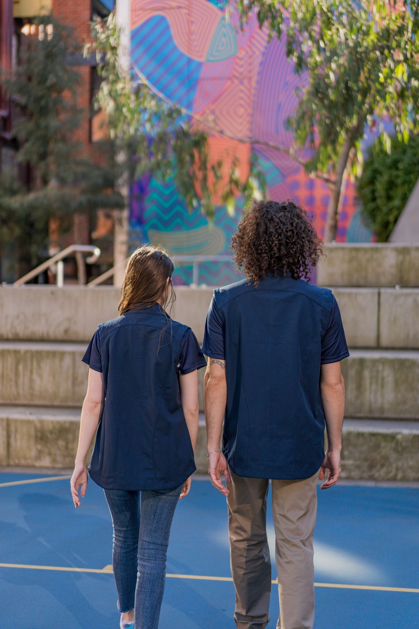 Rear view of the Health, Physical Education & Sport Uniform – Vests worn by a male and a female model standing outdoors on campus. The models are walking side by side in a campus setting. Available at the RMIT Store.