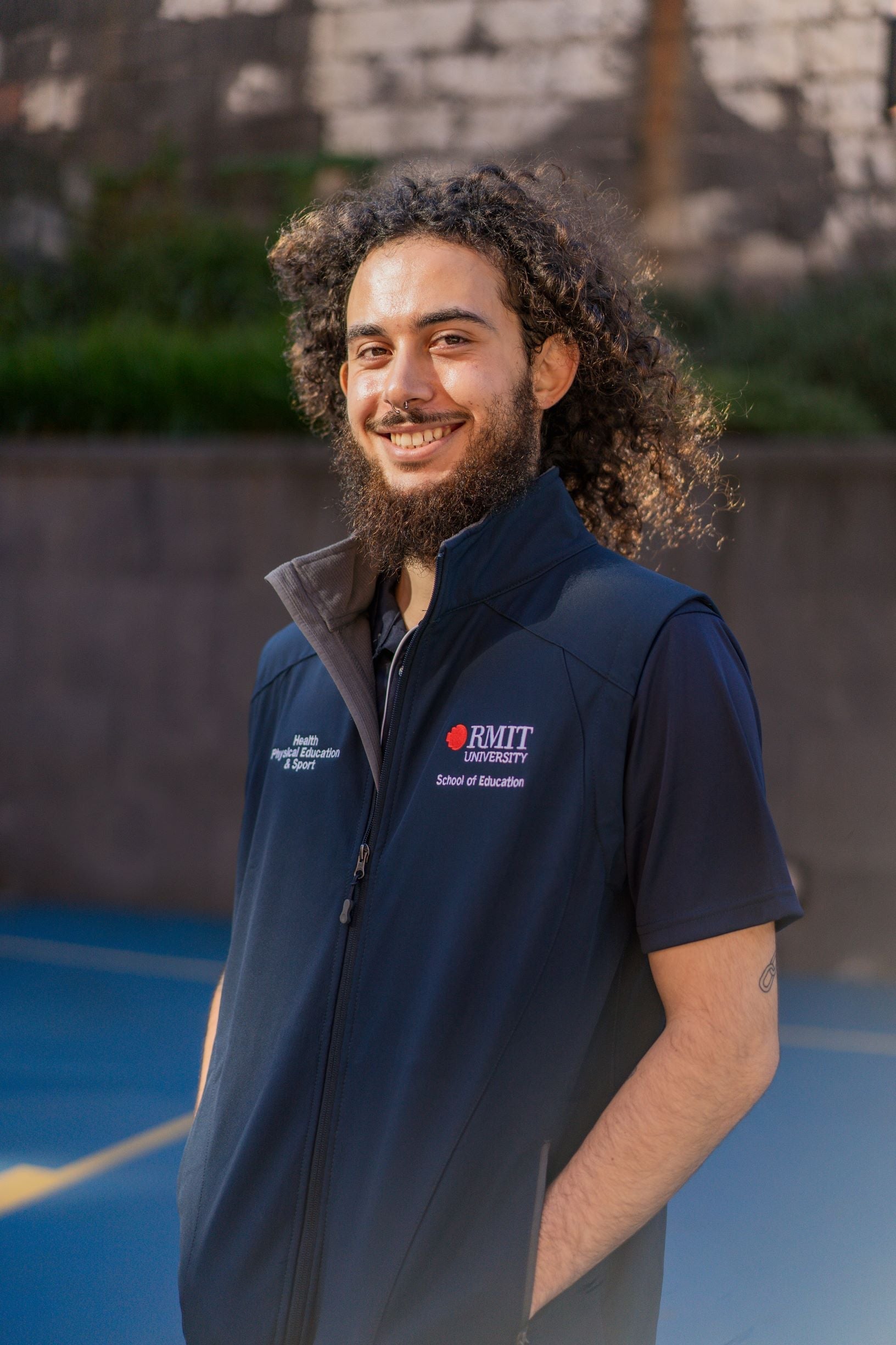 Full front view of the Health, Physical Education & Sport Uniform – Vest worn by a male model standing outdoors on campus with hands in his pockets. The navy polyester vest features white embroidered text reading "Health, Physical Education & Sport" on the right chest, and on the left chest, the RMIT University logo with “School of Education” embroidered underneath. Available at the RMIT Store.