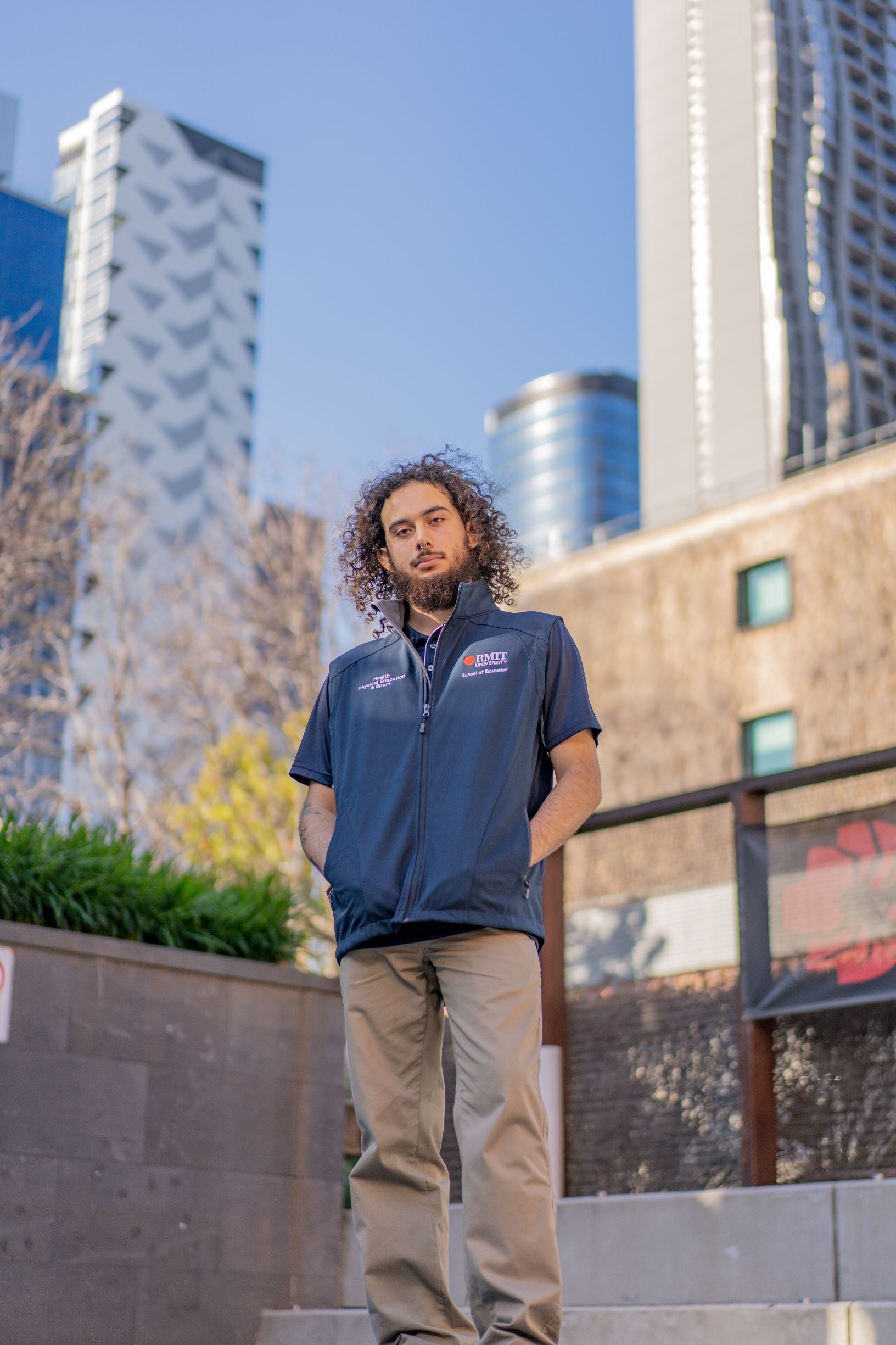 Full front view of the Health, Physical Education & Sport Uniform – Vest worn by a male model looking down toward the camera in an outdoor campus setting. The navy vest features white embroidered text reading "Health, Physical Education & Sport" on the right chest, and the RMIT University logo with "School of Education" underneath on the left chest. Available at the RMIT Store.