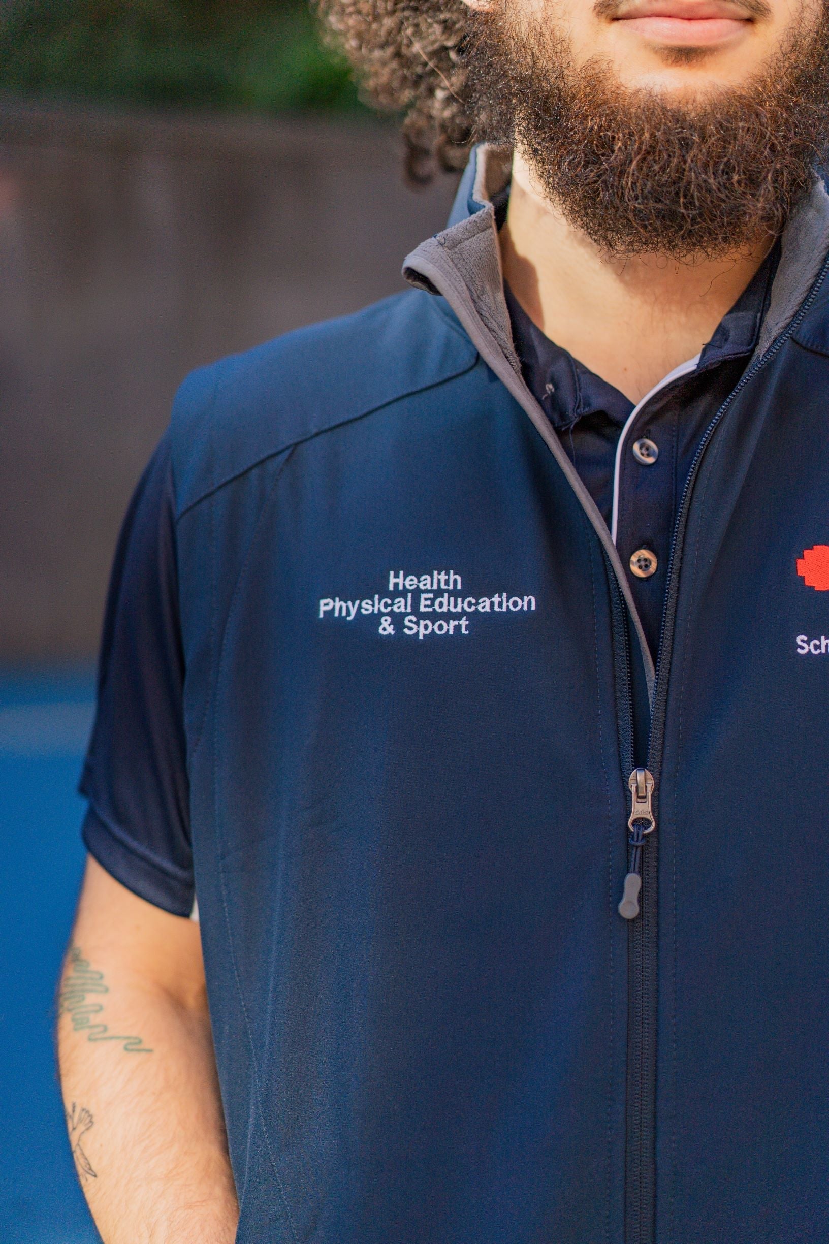 Zoom-in view of the right chest of the Health, Physical Education & Sport Uniform – Vest, featuring white embroidered text that reads "Health, Physical Education & Sport" on navy polyester fabric. The vest is worn by a male model standing outdoors in a campus setting. Available at the RMIT Store.