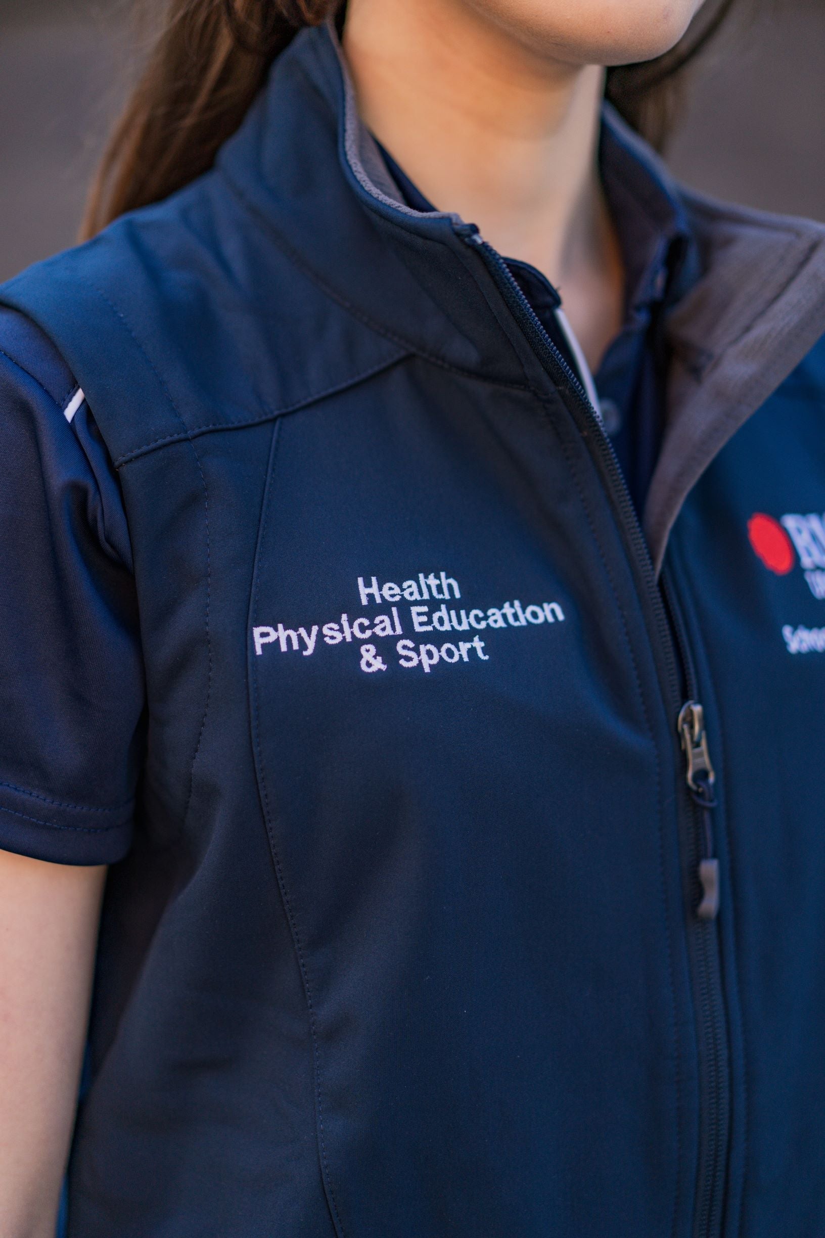 Close-up of the right chest area of a navy Health, Physical Education & Sport Uniform vest worn by a female model, featuring white embroidered text reading “Health, Physical Education & Sport” from the RMIT Store, against an outdoor campus background.