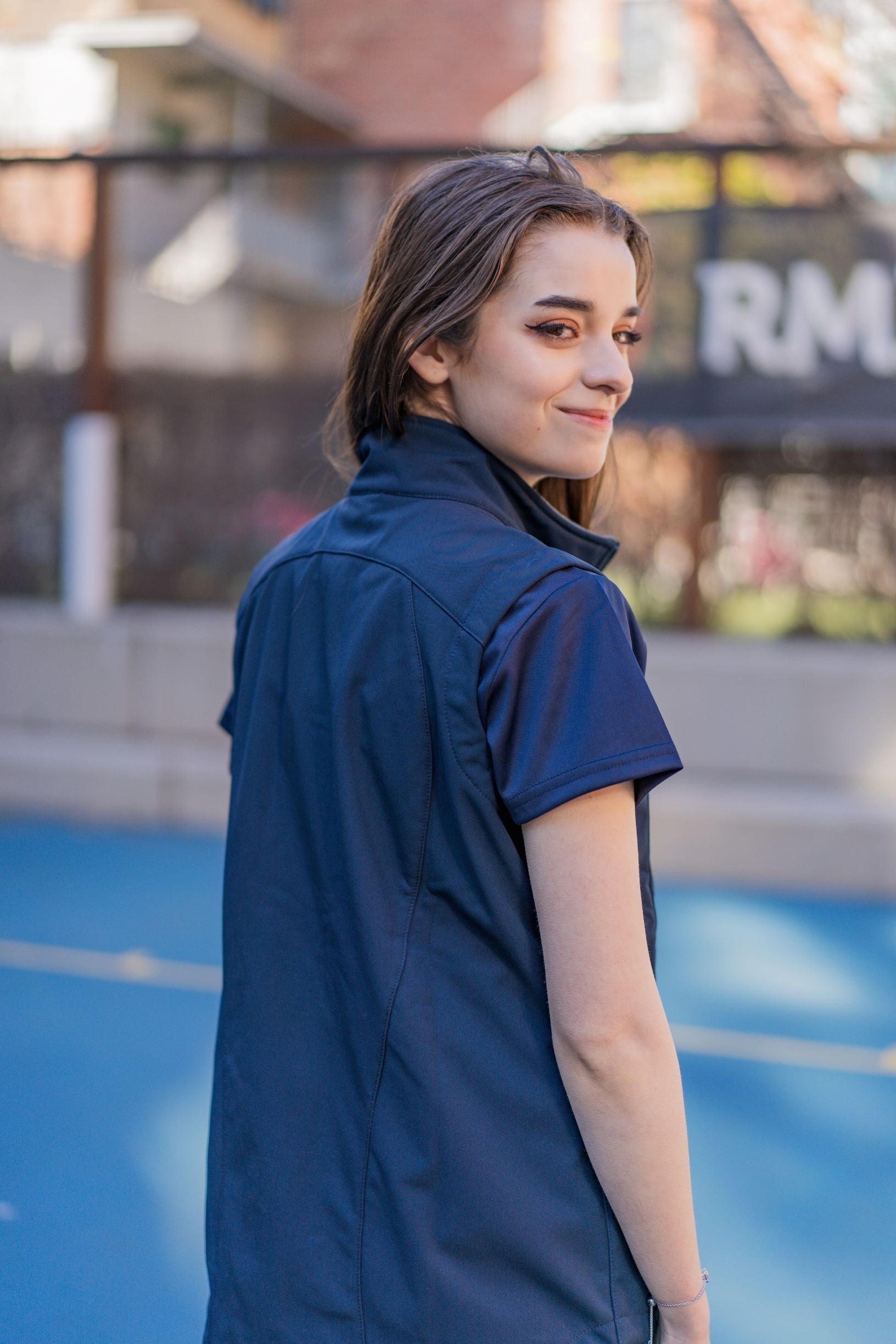 Female model looking back over her shoulder wearing a plain navy Health, Physical Education & Sport Uniform vest from the RMIT Store, outdoors on campus with blurred background.