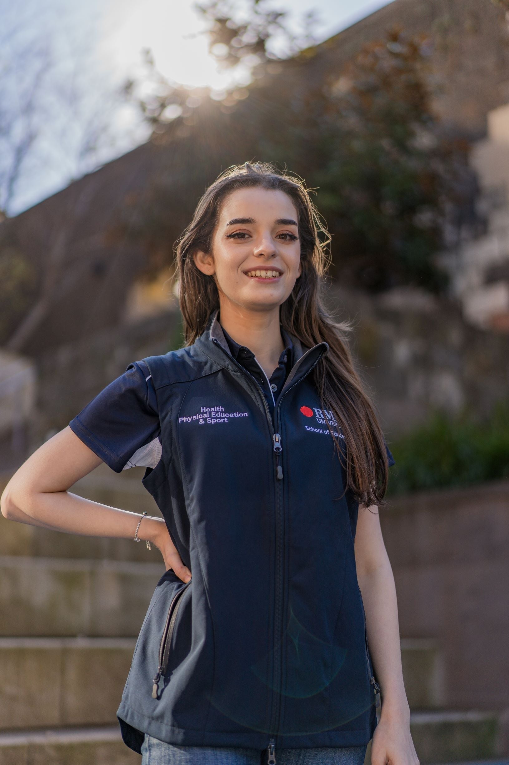 Female model wearing the navy Health, Physical Education & Sport Uniform – Vest with white embroidered “Health, Physical Education & Sport” text on the right chest, one hand on her waist, standing outdoors on campus; the vest is worn over a matching polo shirt. Available at RMIT Store.
